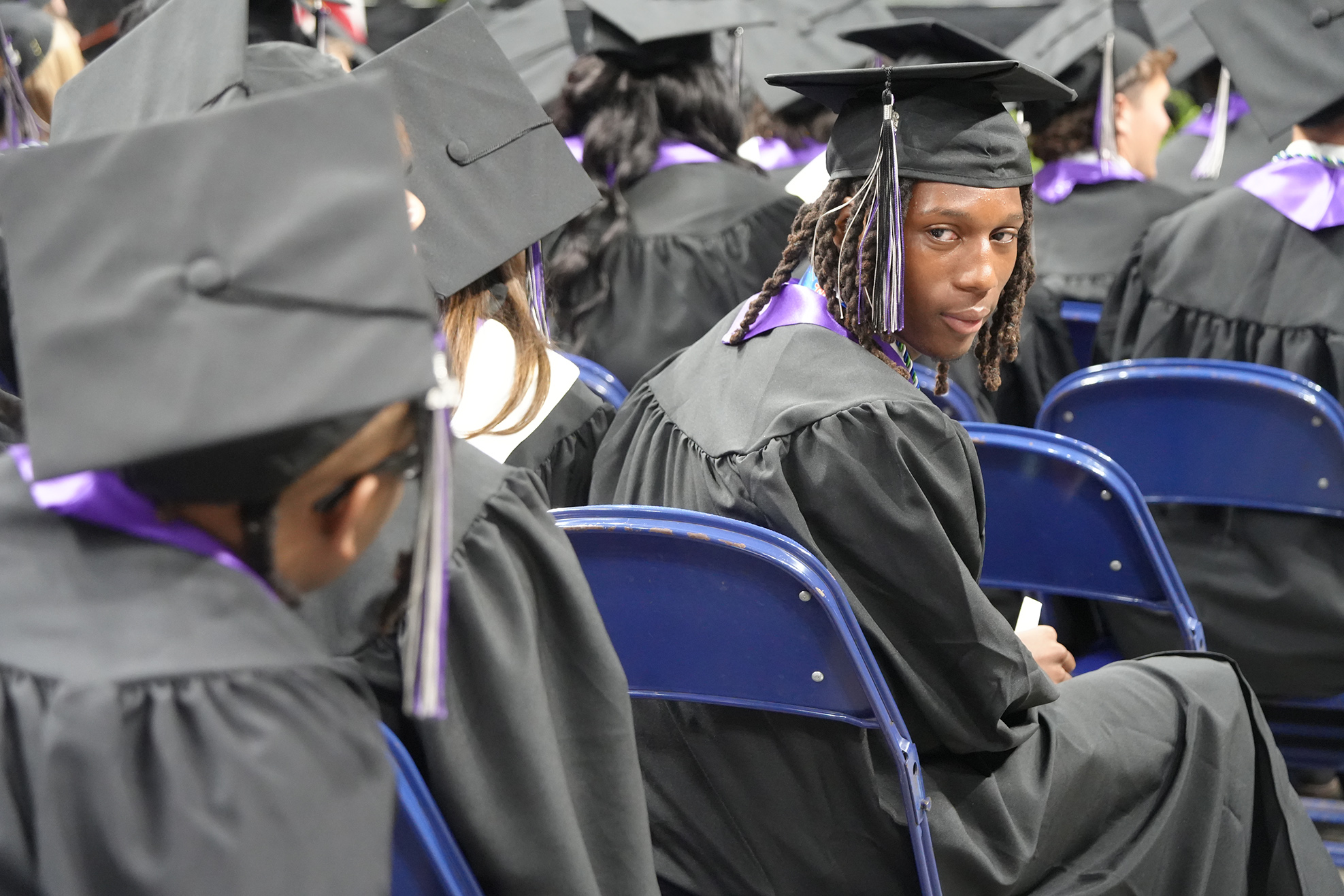 Graduating students in black gowns and caps are seated in blue chairs at a ceremony.