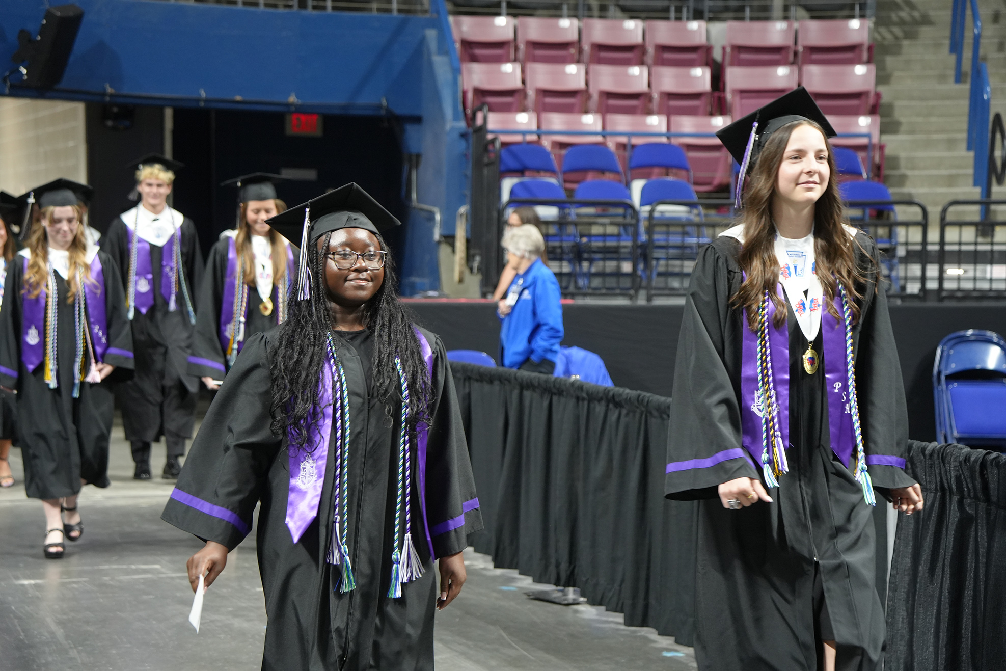 Graduates in black gowns and caps walk across a stage, with empty seats and a blue wall in the background.