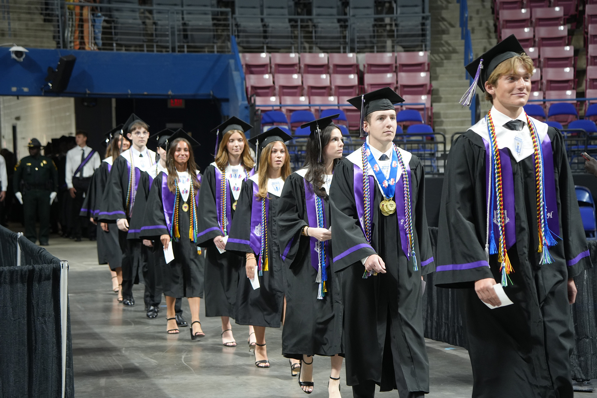 A group of students in graduation attire walks on stage with a large, partially empty stadium in the background.