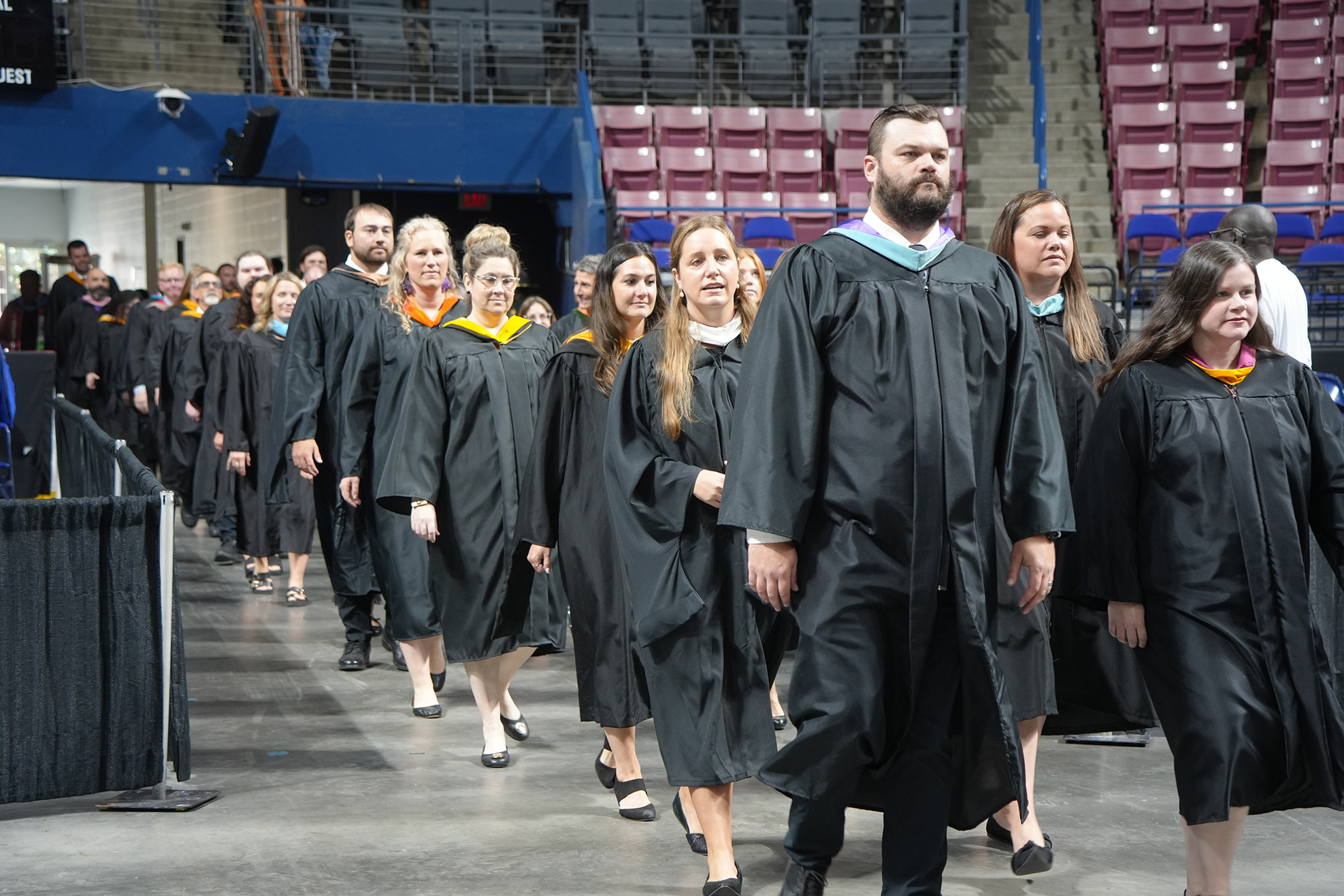 Graduates in black gowns walk in a line, possibly for a ceremony. Behind them is a stadium with empty seats.