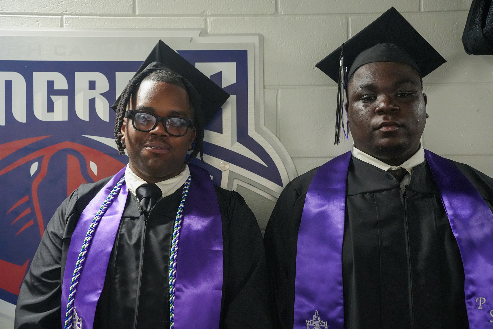 Two people in academic gowns and caps pose next to a wall with a logo.