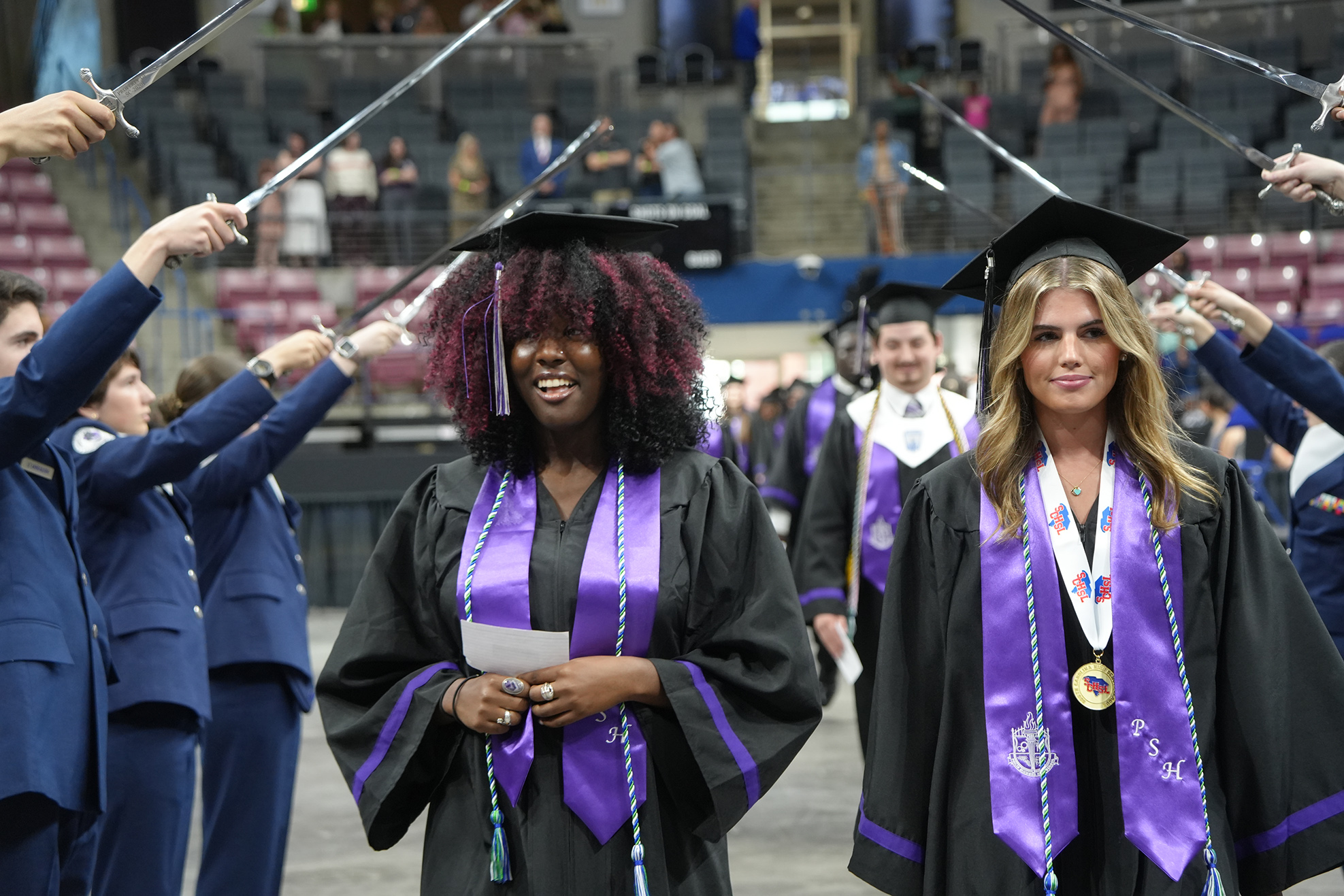 Graduates in caps and gowns walk in a line at a ceremony. One holds a paper and smiles.