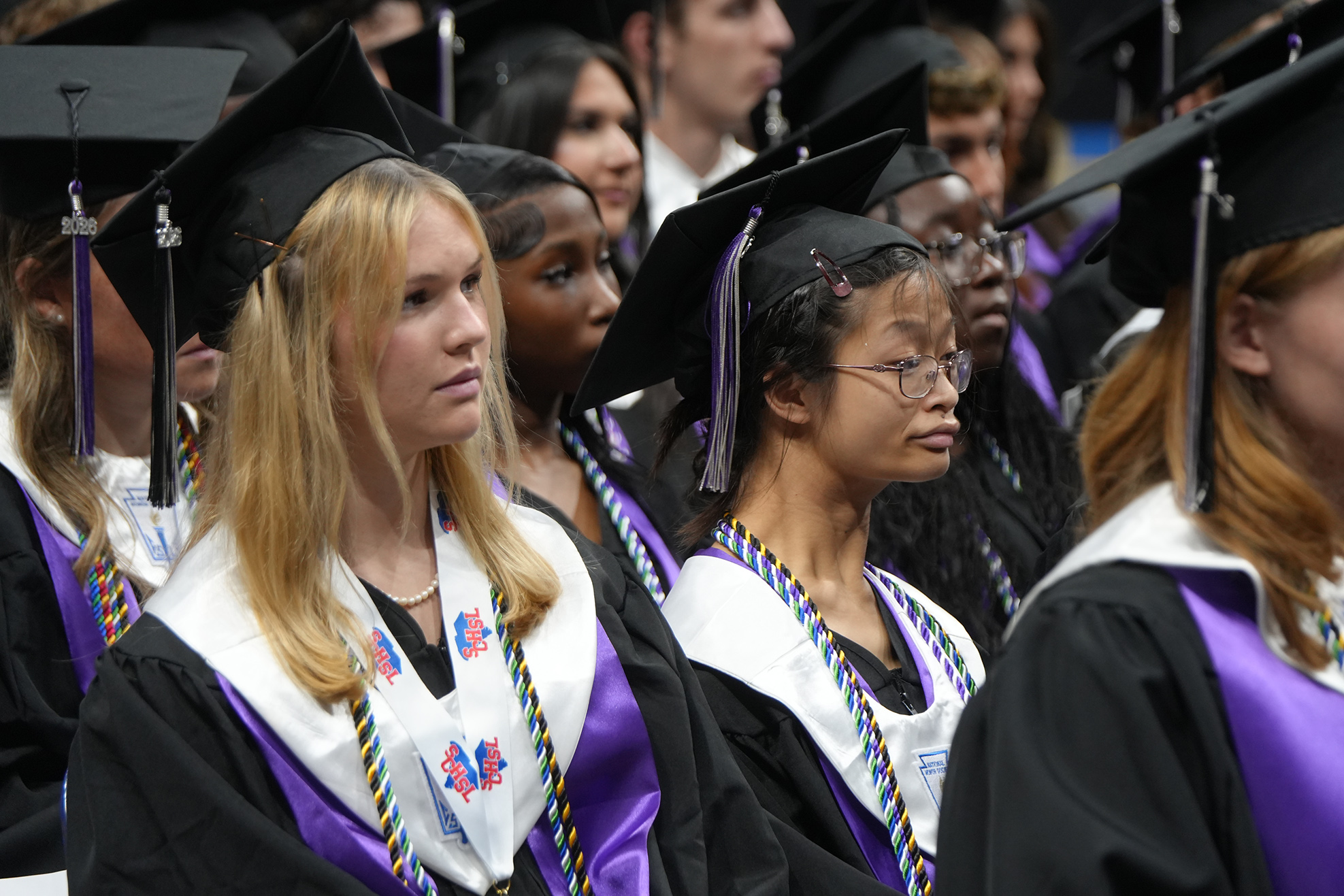 A group of graduates wearing black gowns and purple stoles are lined up. Their tassels are black.