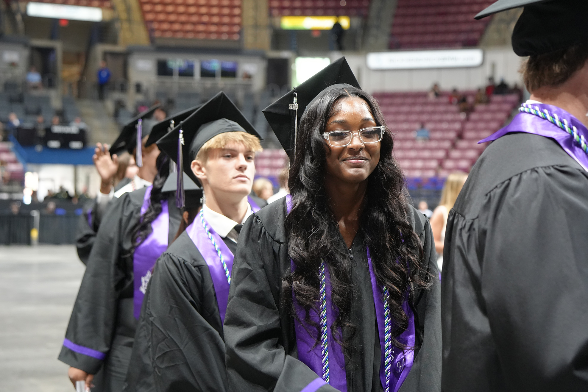 A woman in a cap and gown stands with others at a ceremony in an indoor arena.