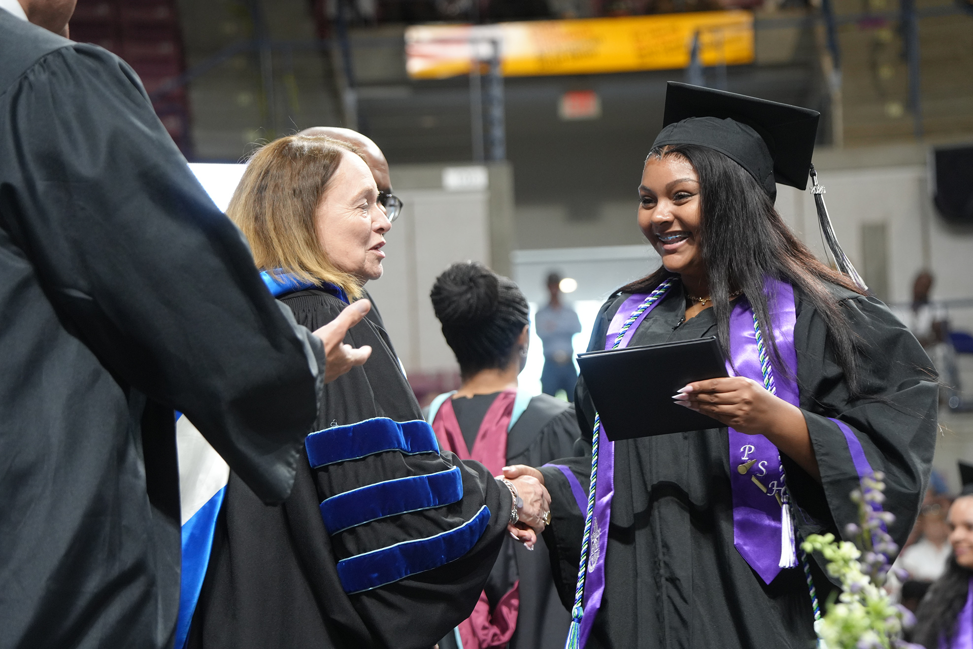 A woman in a black gown and mortarboard shakes hands with a faculty member at a graduation ceremony.