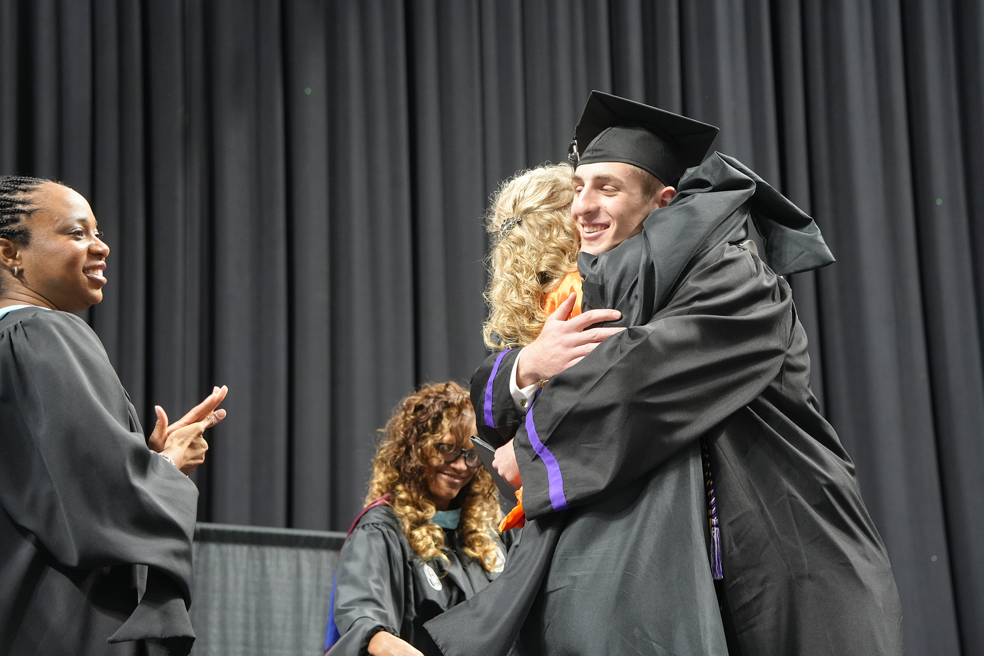 Two graduates in black gowns and caps hug on stage. A smiling woman stands to the left, while another graduate watches behind them. Black curtains are in the background.