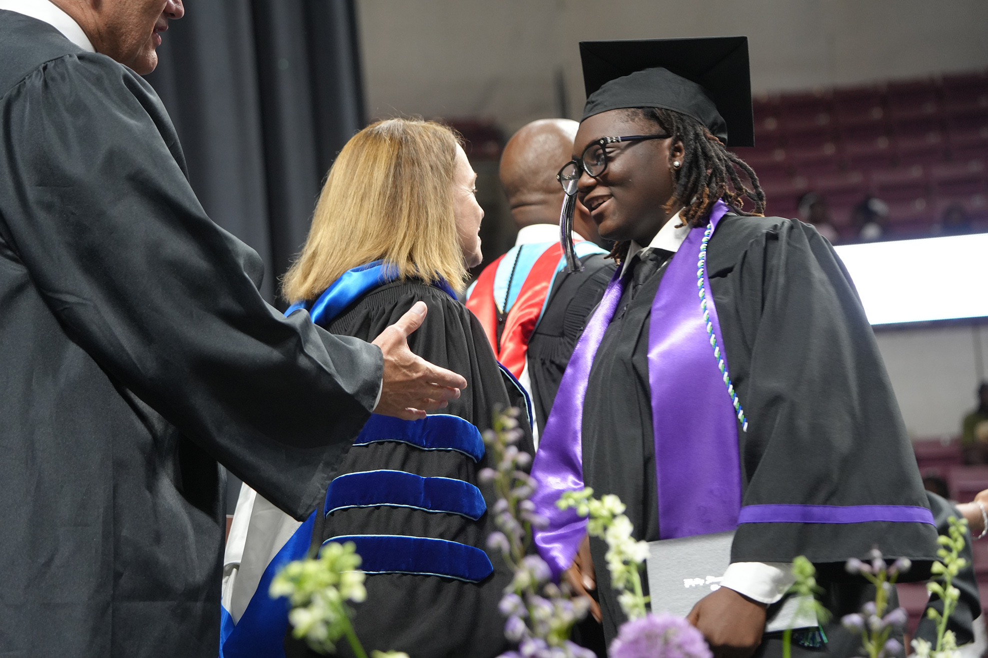 A man and a woman in academic attire, with the man giving a handshake to the woman.