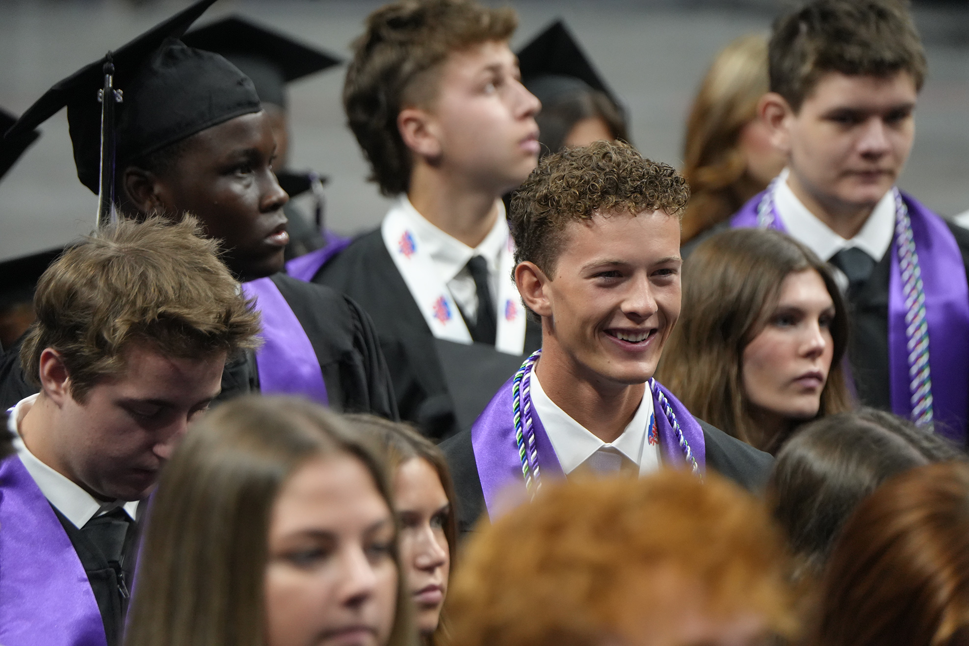 A crowd of students in graduation gowns and caps stand together, facing forward. Some have their heads turned.