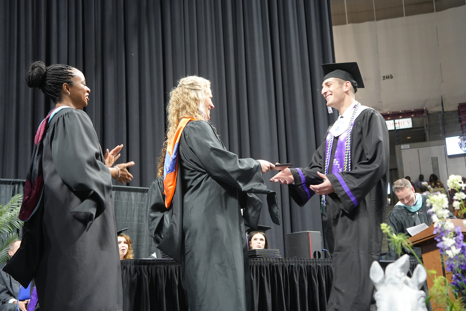 A woman hands a diploma to a man in front of an audience; both wear graduation gowns.