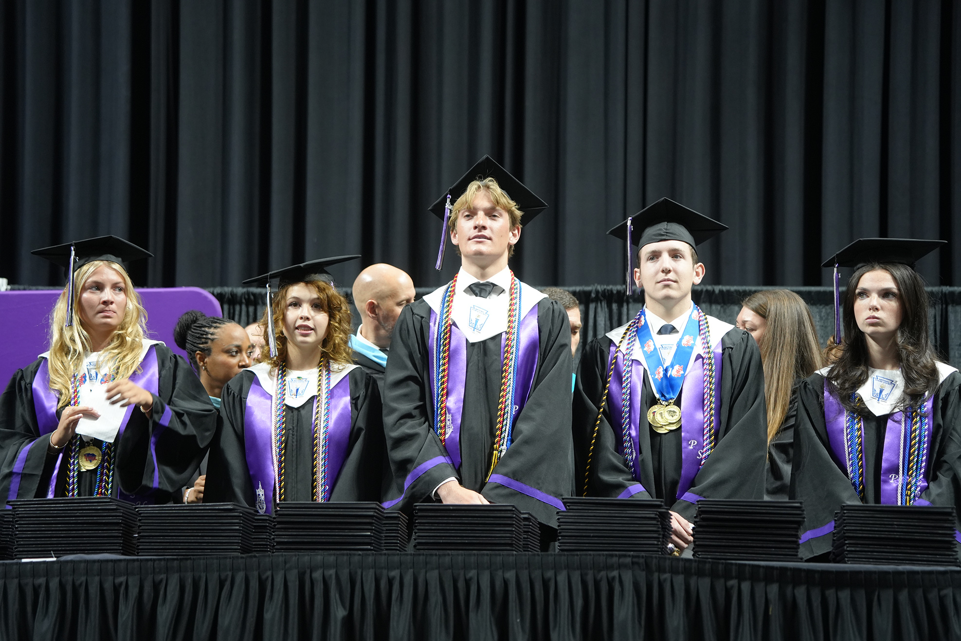 A group of graduates in black gowns and caps stand on a stage with podiums in front.