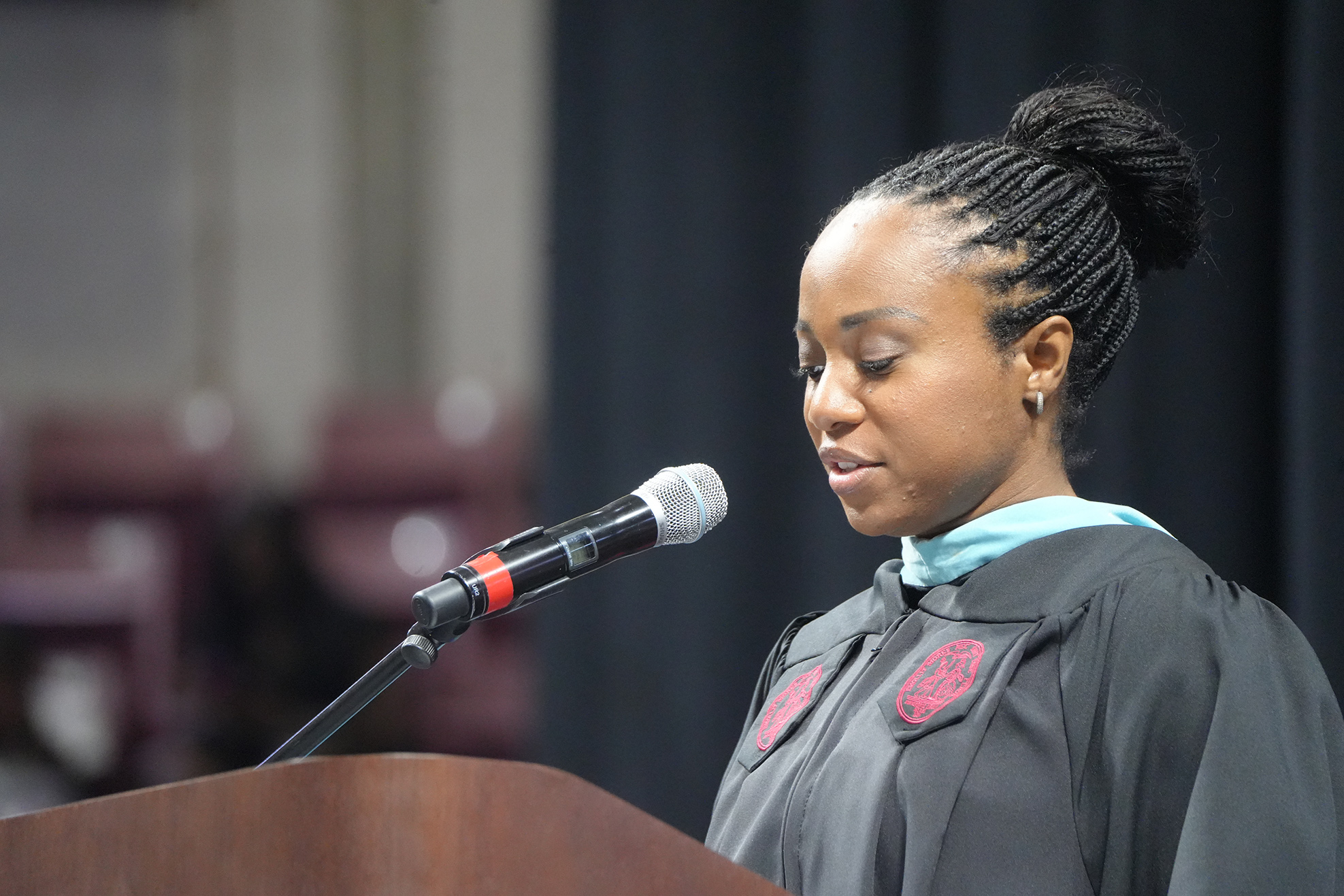 A woman wearing a black gown and blue stole stands at a podium with a microphone.