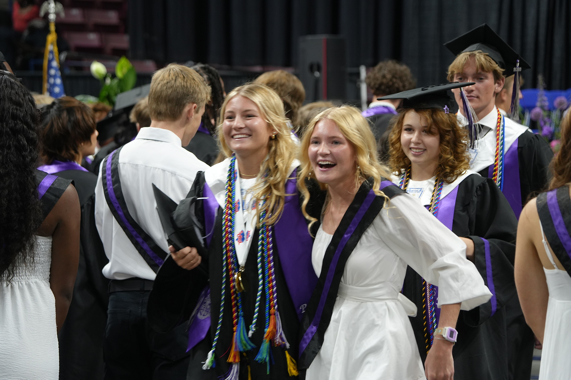 Group of people in caps and gowns, some smiling. Two women, one in white, both with graduation cords, stand close.