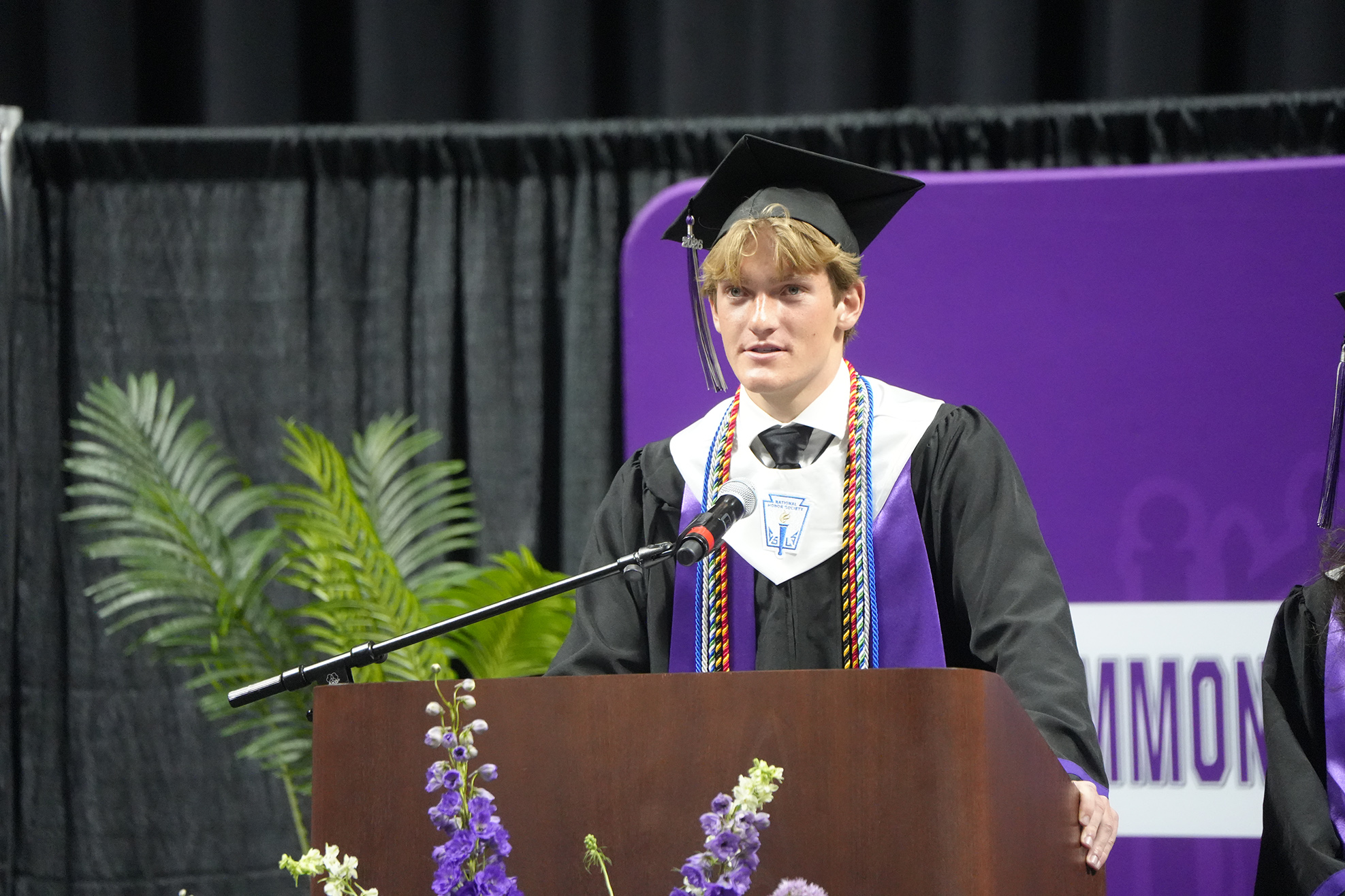 A person in a cap and gown stands at a podium, with a microphone, a plant, and purple flowers.