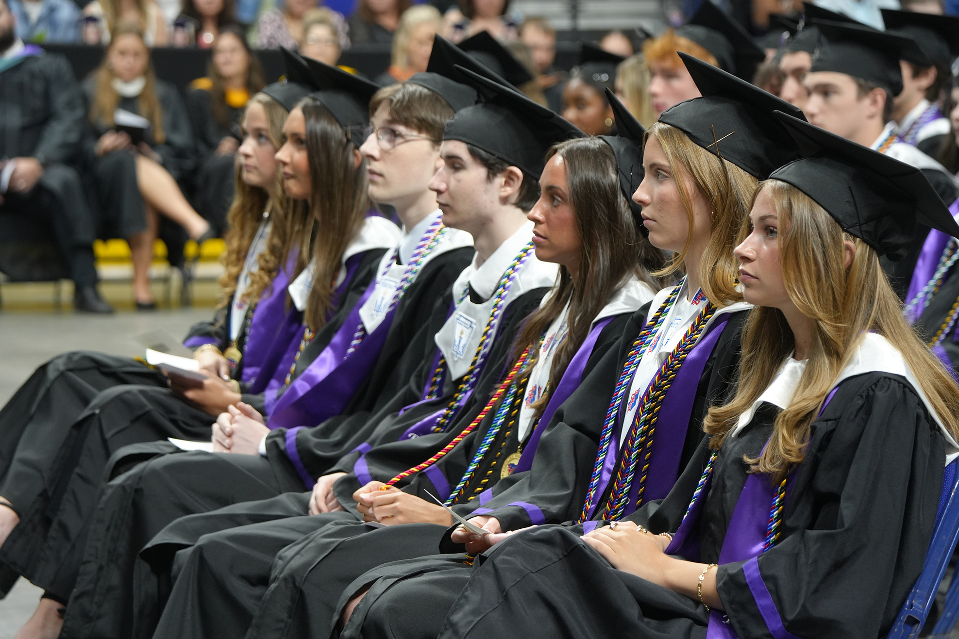 Graduates in caps and gowns sit in a row, some with hands on laps, in a stadium.