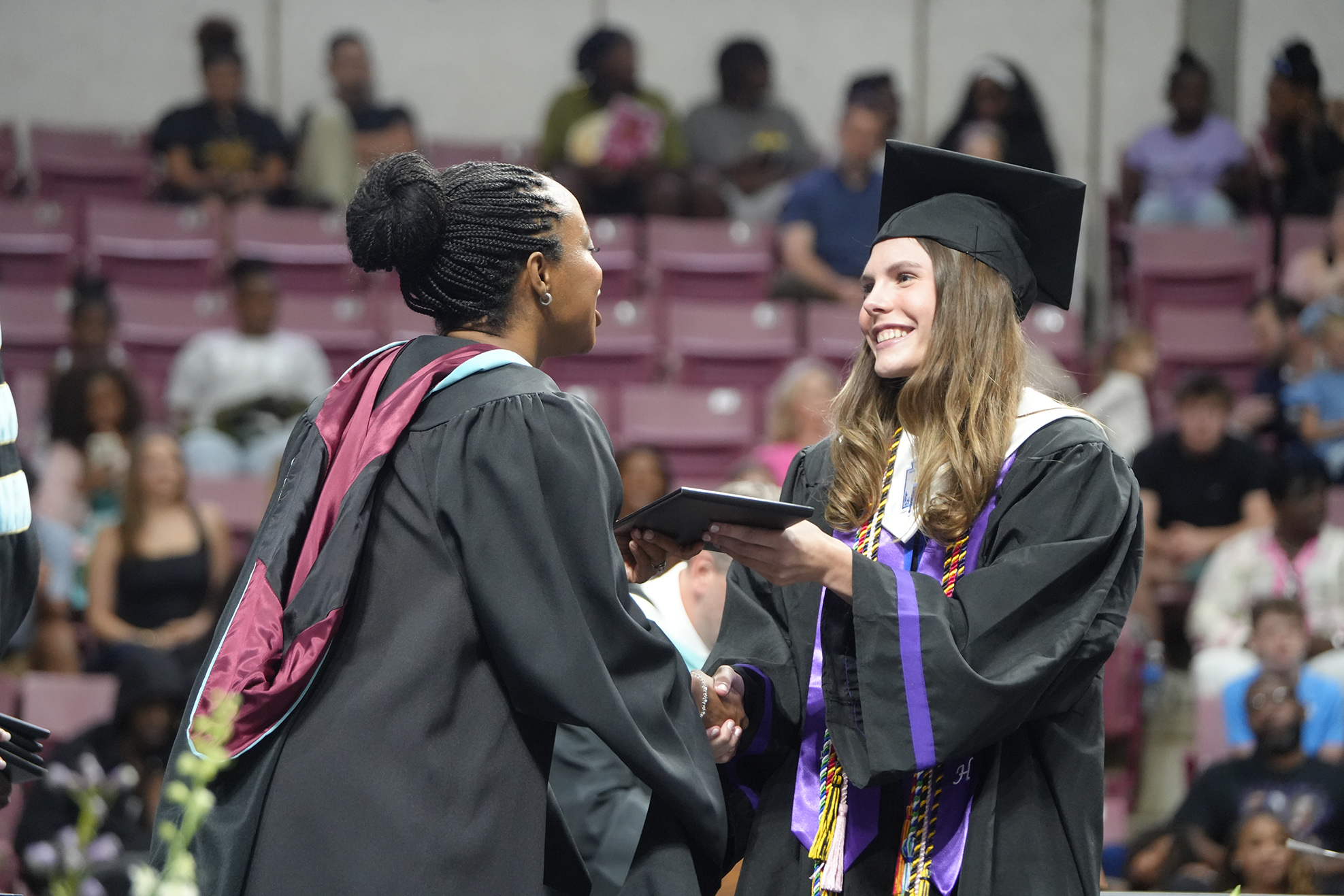 Two women in caps and gowns shake hands in front of an audience in a stadium.