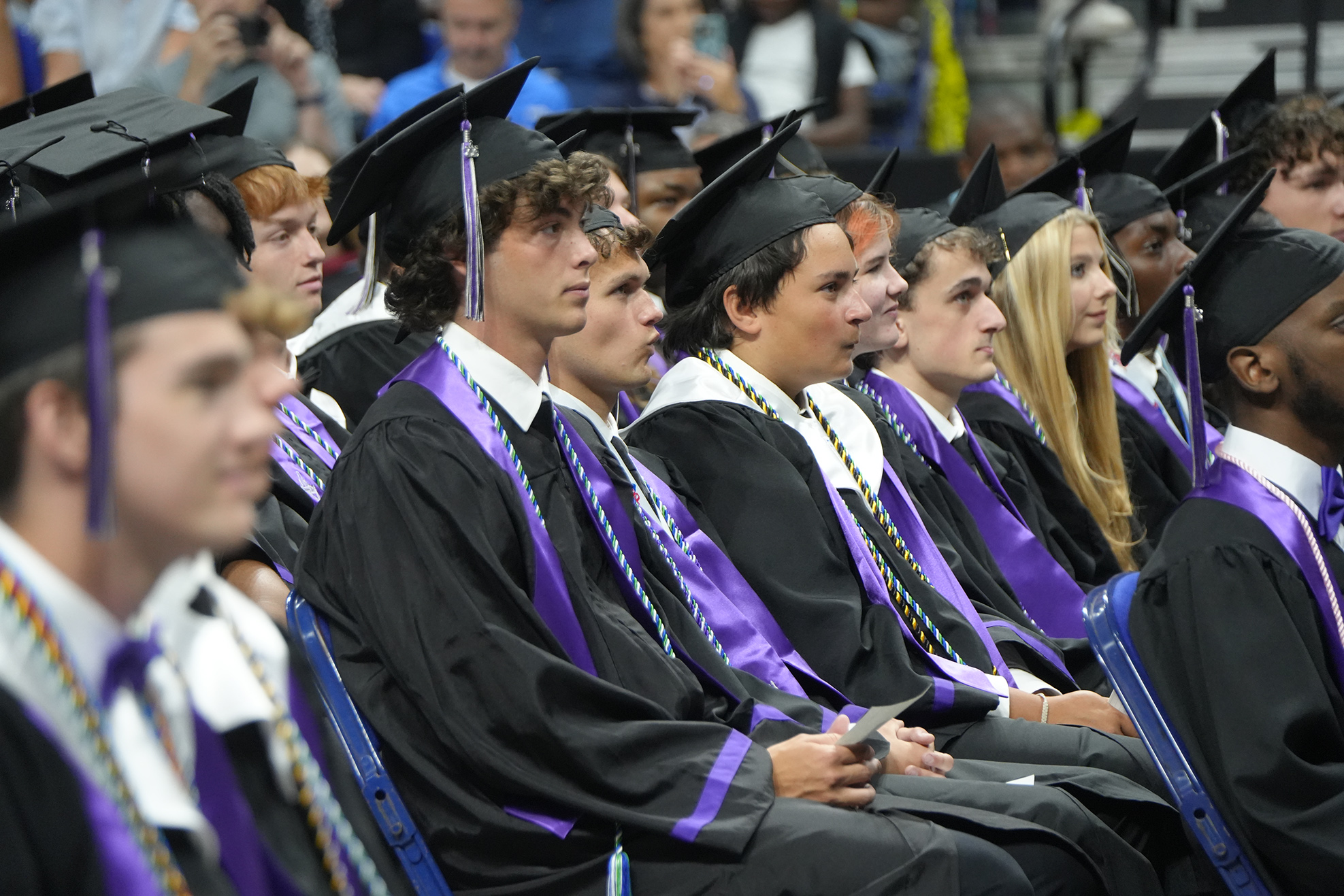 A group of people wearing black graduation gowns and purple stoles sit in rows at an event.