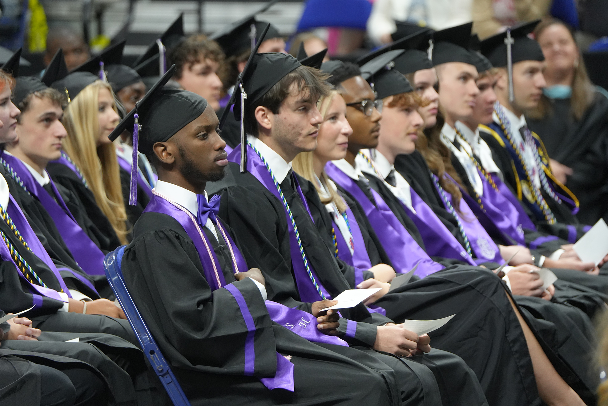 Graduates in black and purple gowns and caps sit in rows at an indoor ceremony.