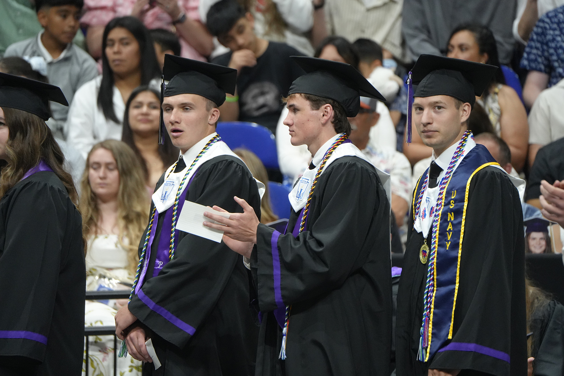 Four individuals in academic regalia, wearing caps and gowns, stand in a row during an outdoor event.