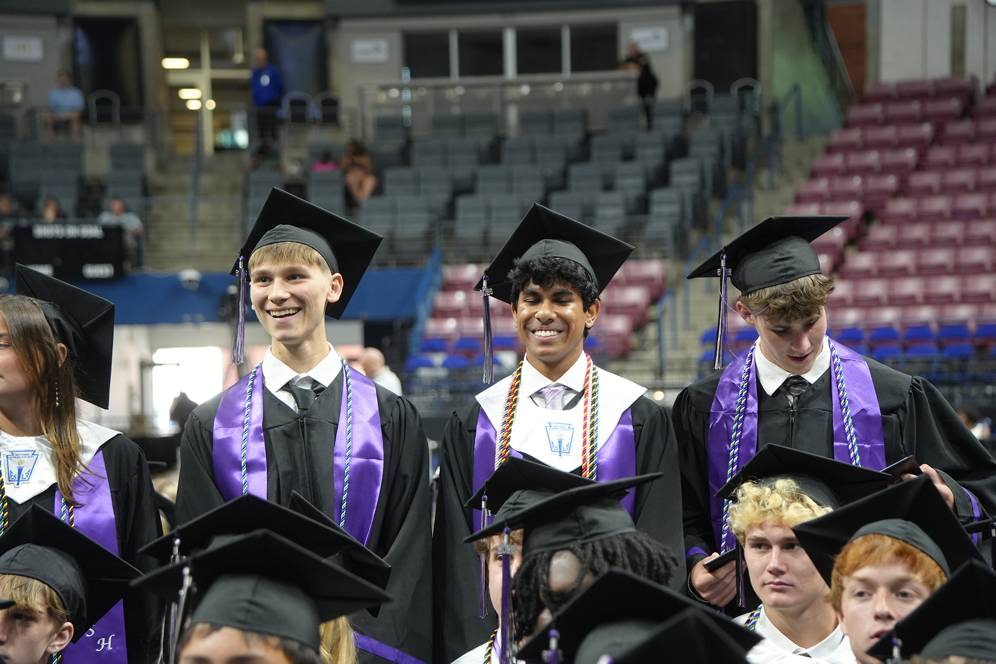 Graduates in caps and gowns stand in an auditorium with empty blue and red seats in the background.