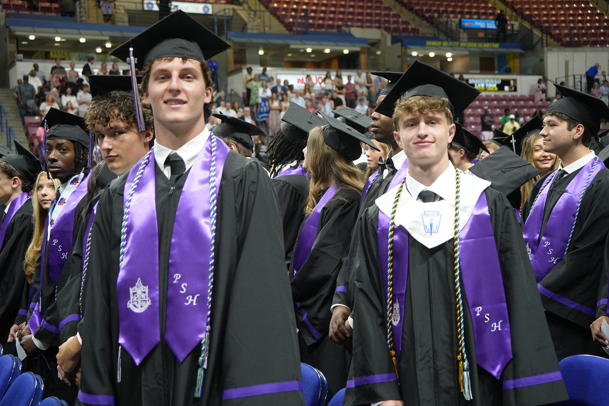 Several young adults wearing graduation attire stand in a line. They wear caps and gowns, and some have sashes across their chests.