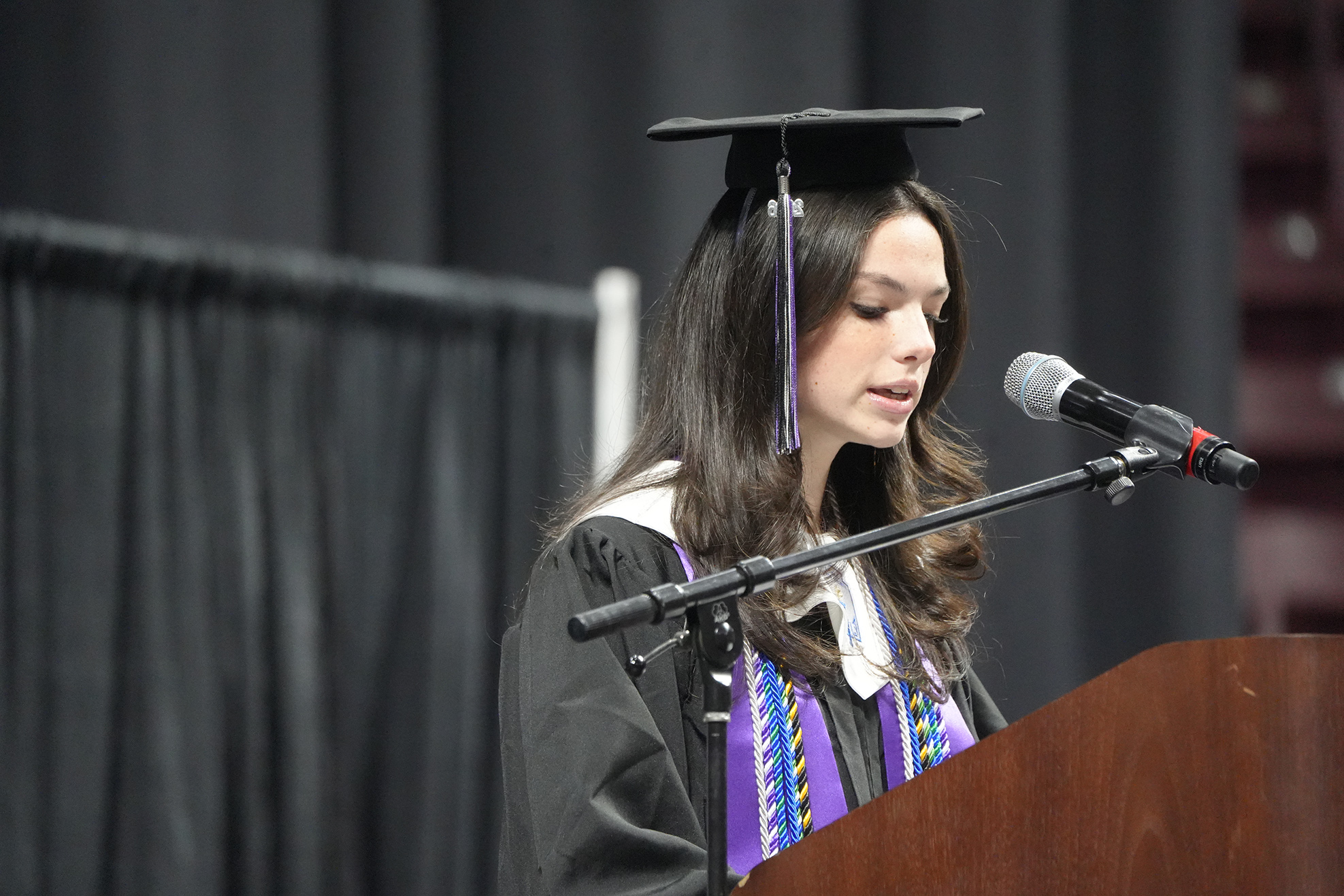A person in a graduation gown, speaking into a microphone at a podium. Behind them is a dark background with curtains.