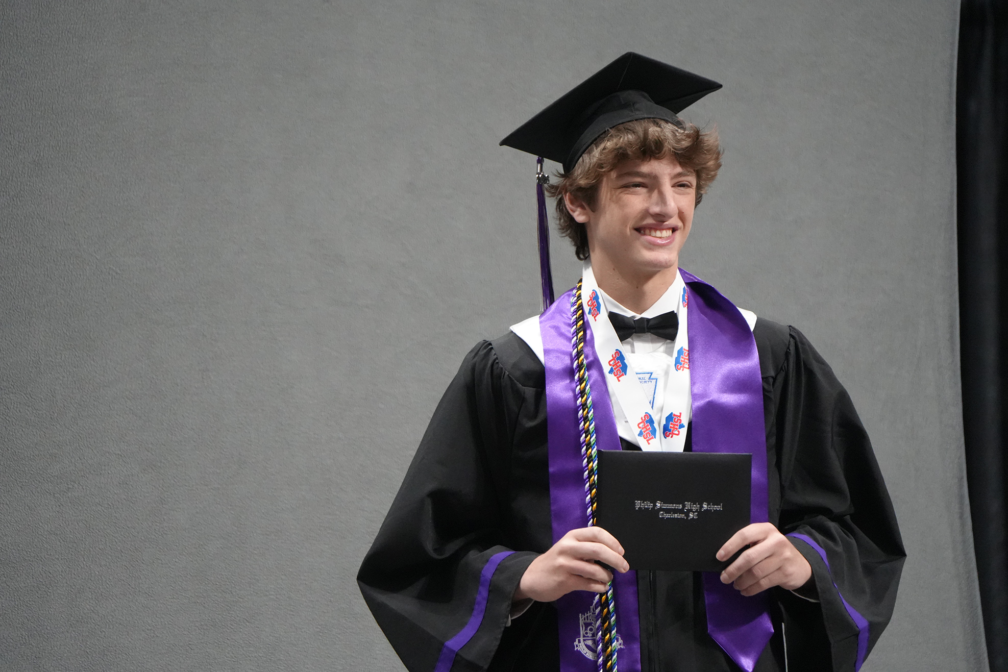 A person in a graduation cap and gown holds a diploma. The gown is black with a purple sash.