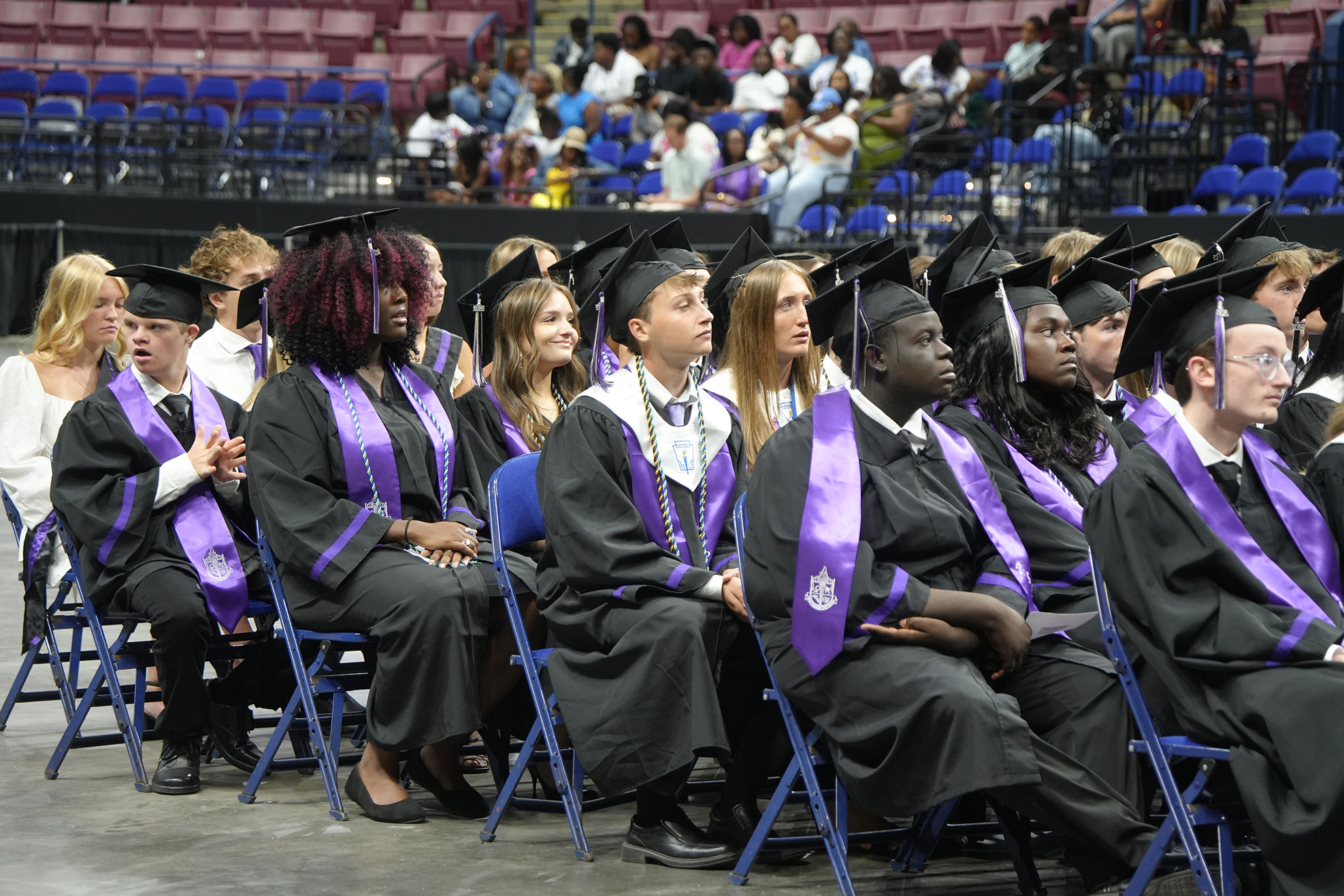 A group of people wearing caps and gowns, seated in blue chairs at an event.