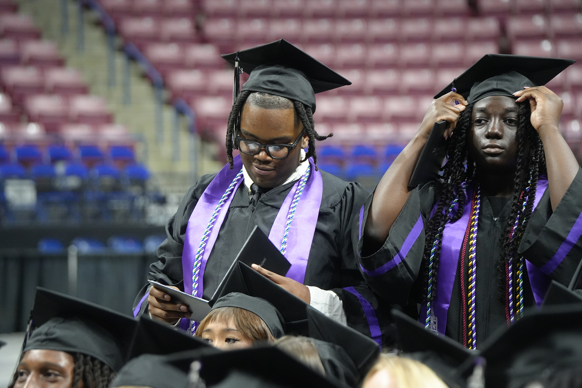 People in graduation gowns and caps, standing in a stadium. One holds a book, another holds their head.