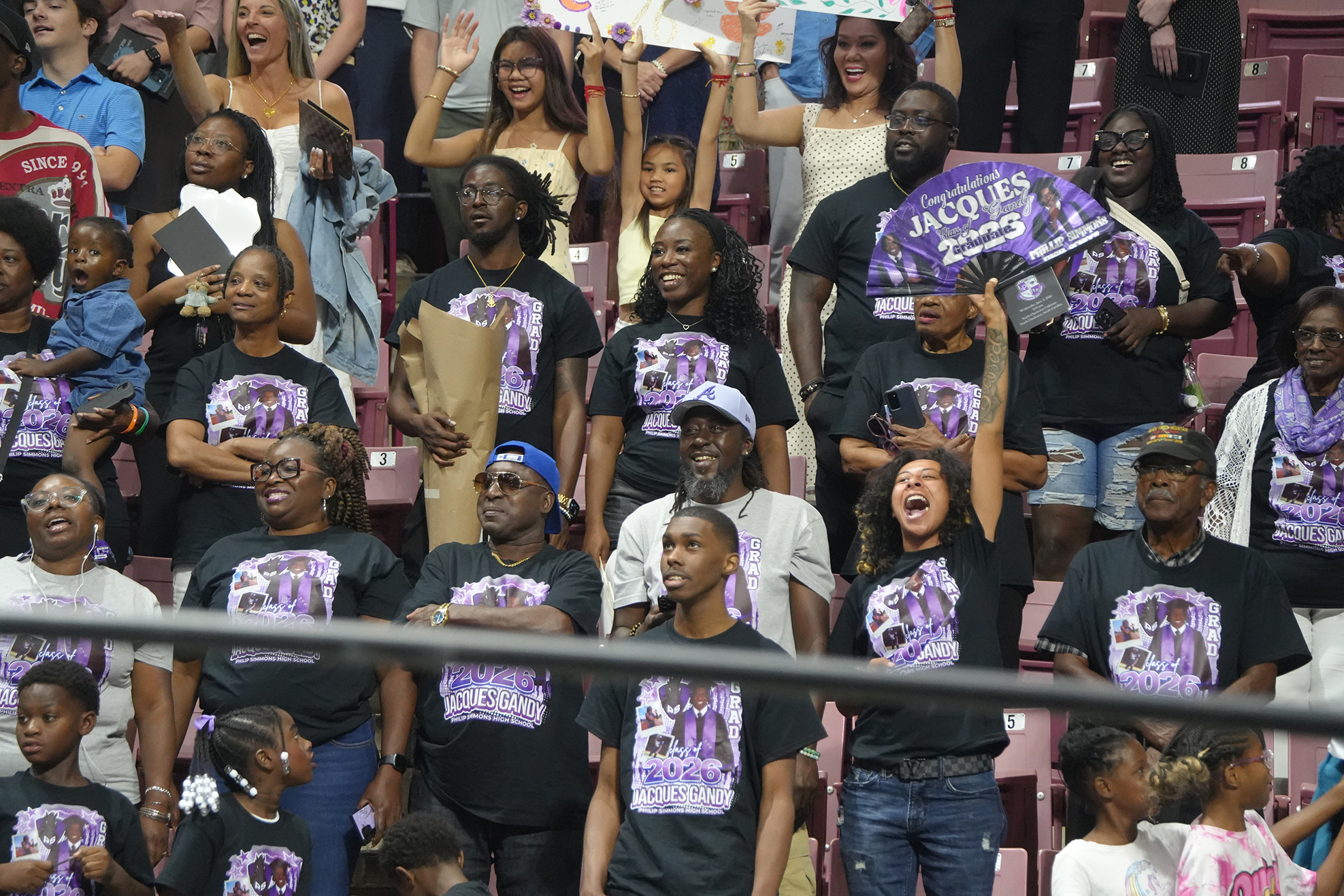 Spectators at a sports event, wearing matching shirts, some with raised hands and others holding signs.