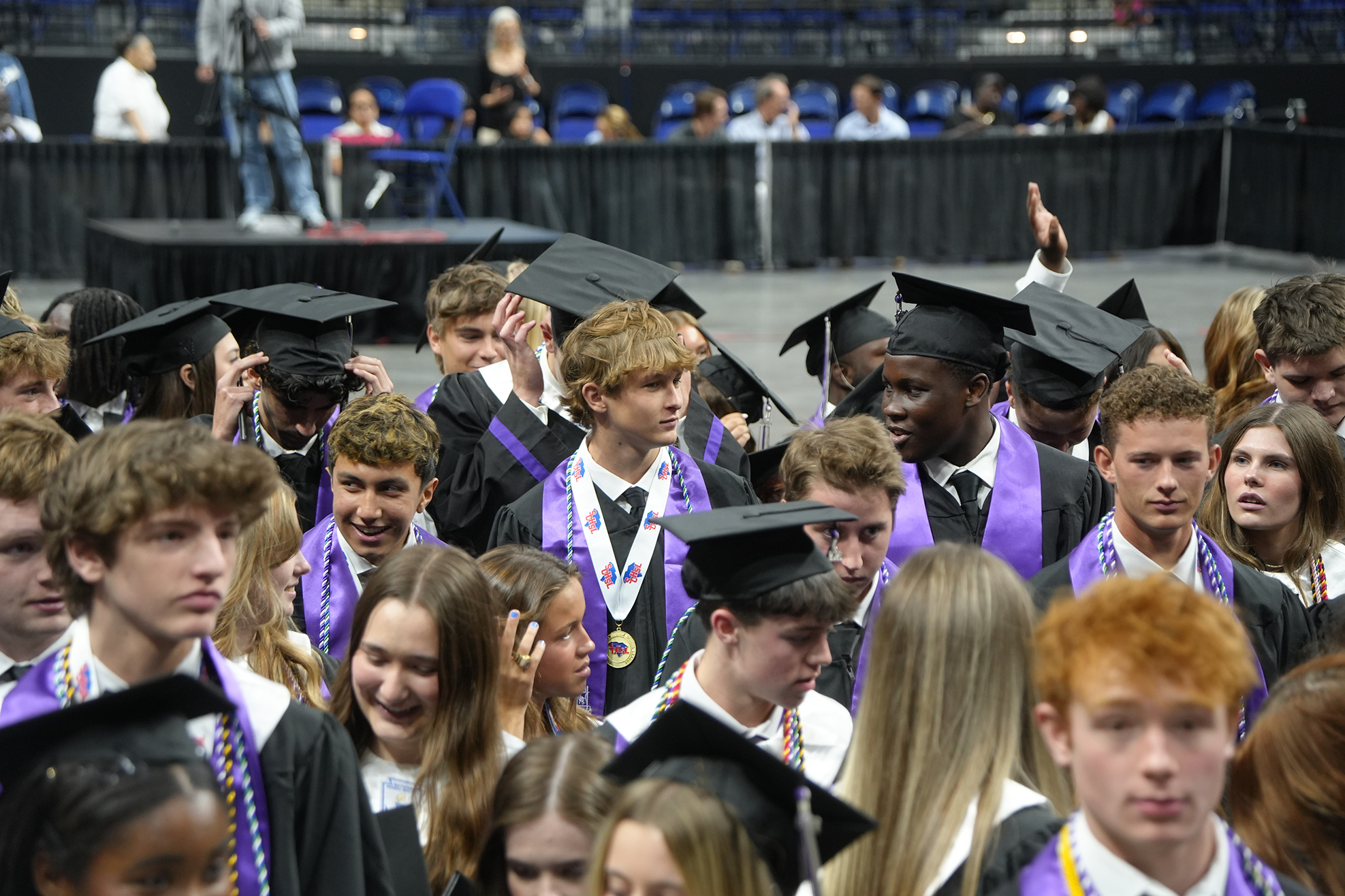Group of students in caps and gowns, some raising their caps, in a large auditorium.