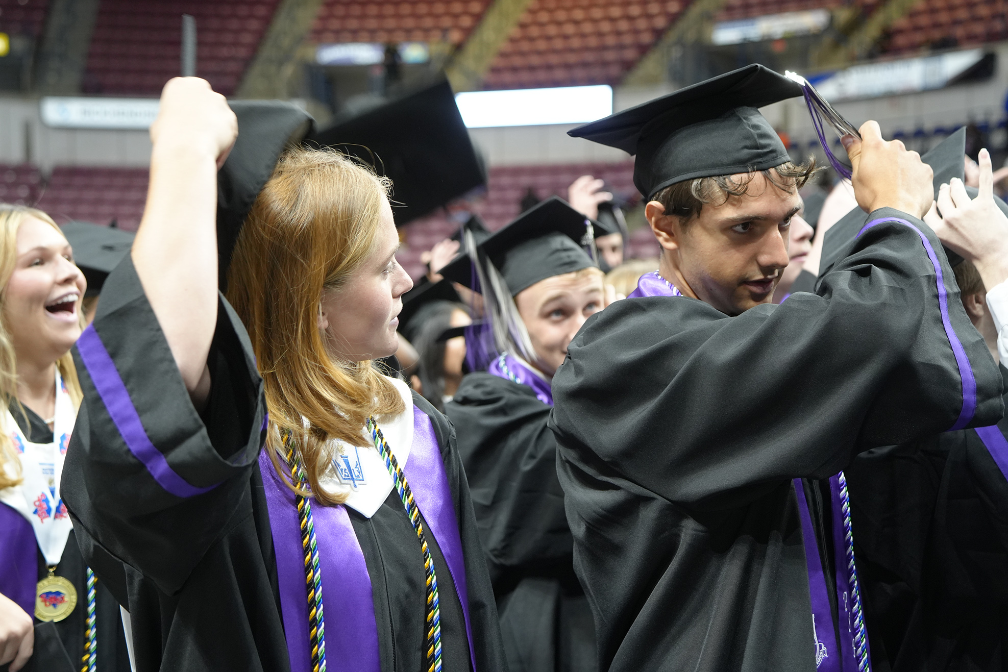Graduates in black and purple gowns and caps stand in a stadium. Some raise their caps.