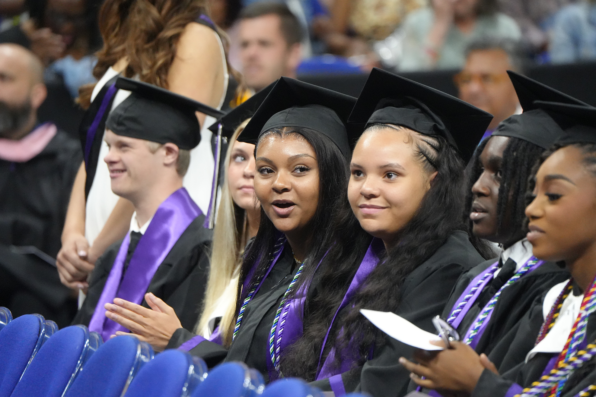 Graduates in black gowns and hats sit in a row, facing front, clapping. A woman with a ponytail is in the middle.