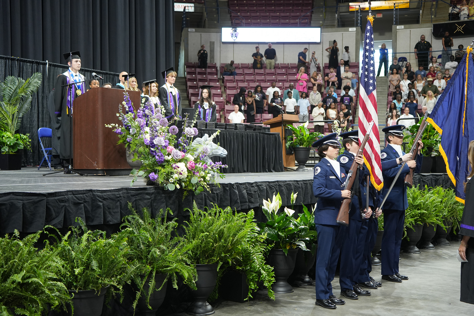 A graduation ceremony stage with a podium, flowers, and military personnel holding flags in front of an audience.