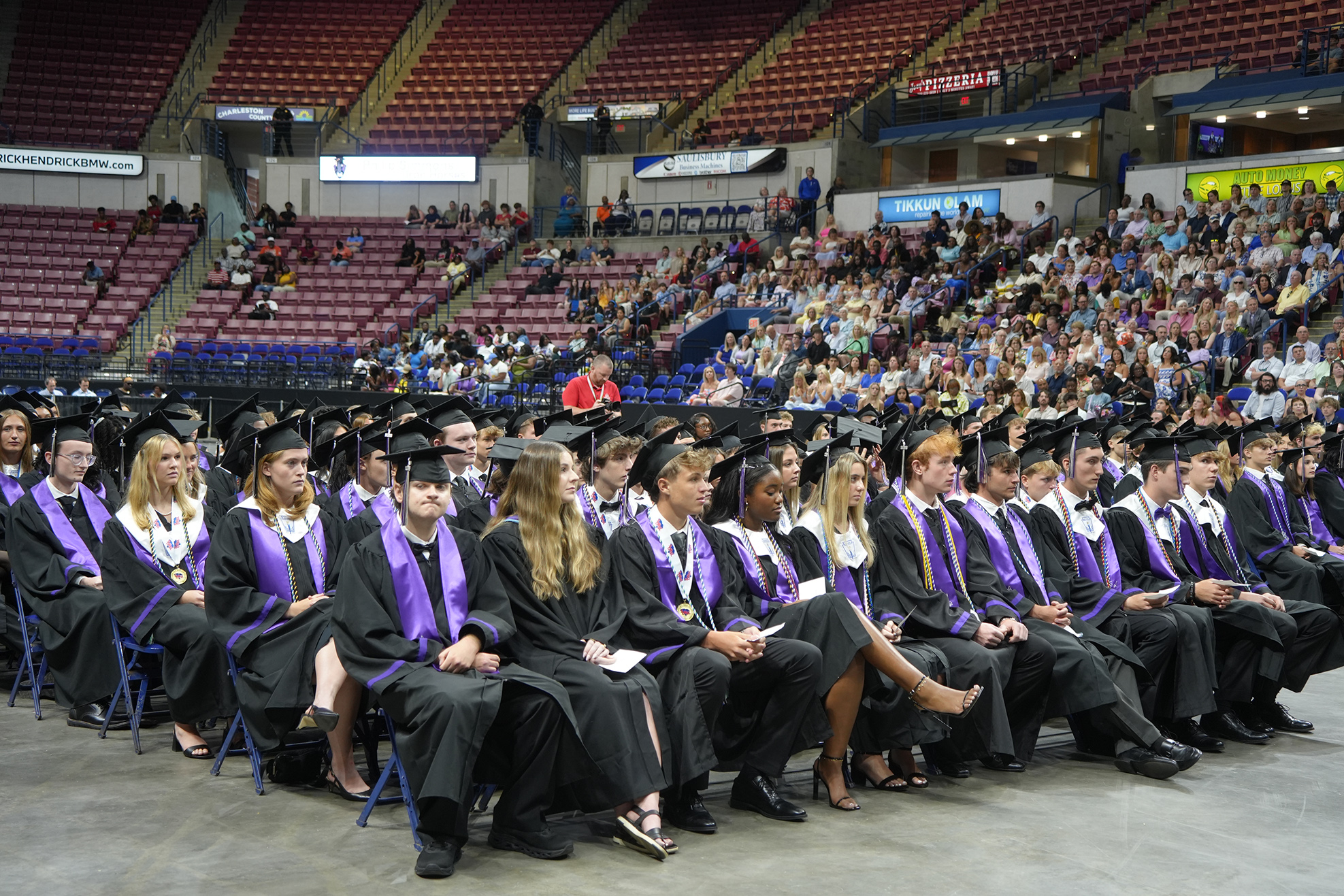 Graduates in caps and gowns sit in rows in an arena. Behind them are rows of empty seats and a large crowd.