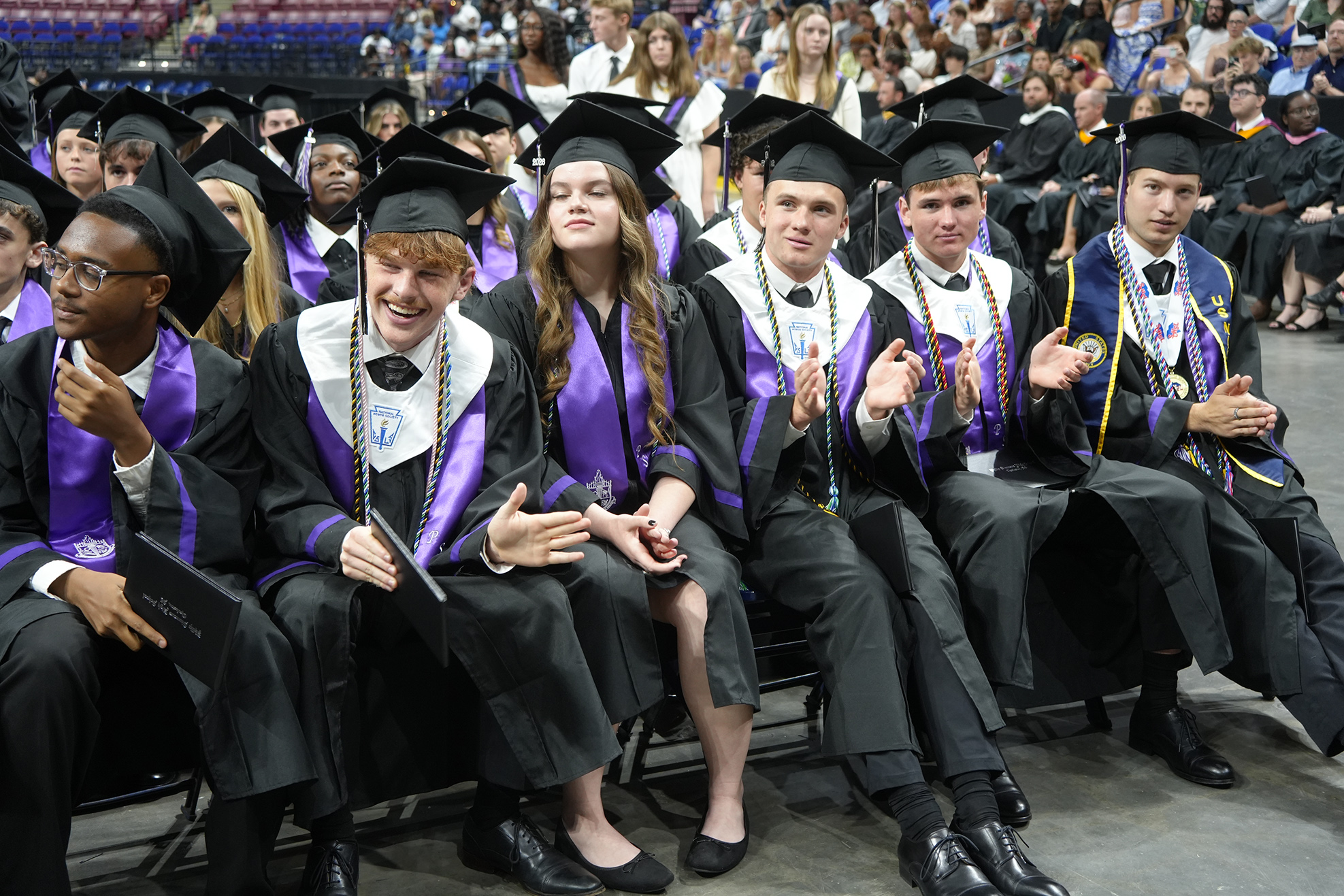 Several people in black and purple graduation gowns and caps sitting in chairs, some clapping.