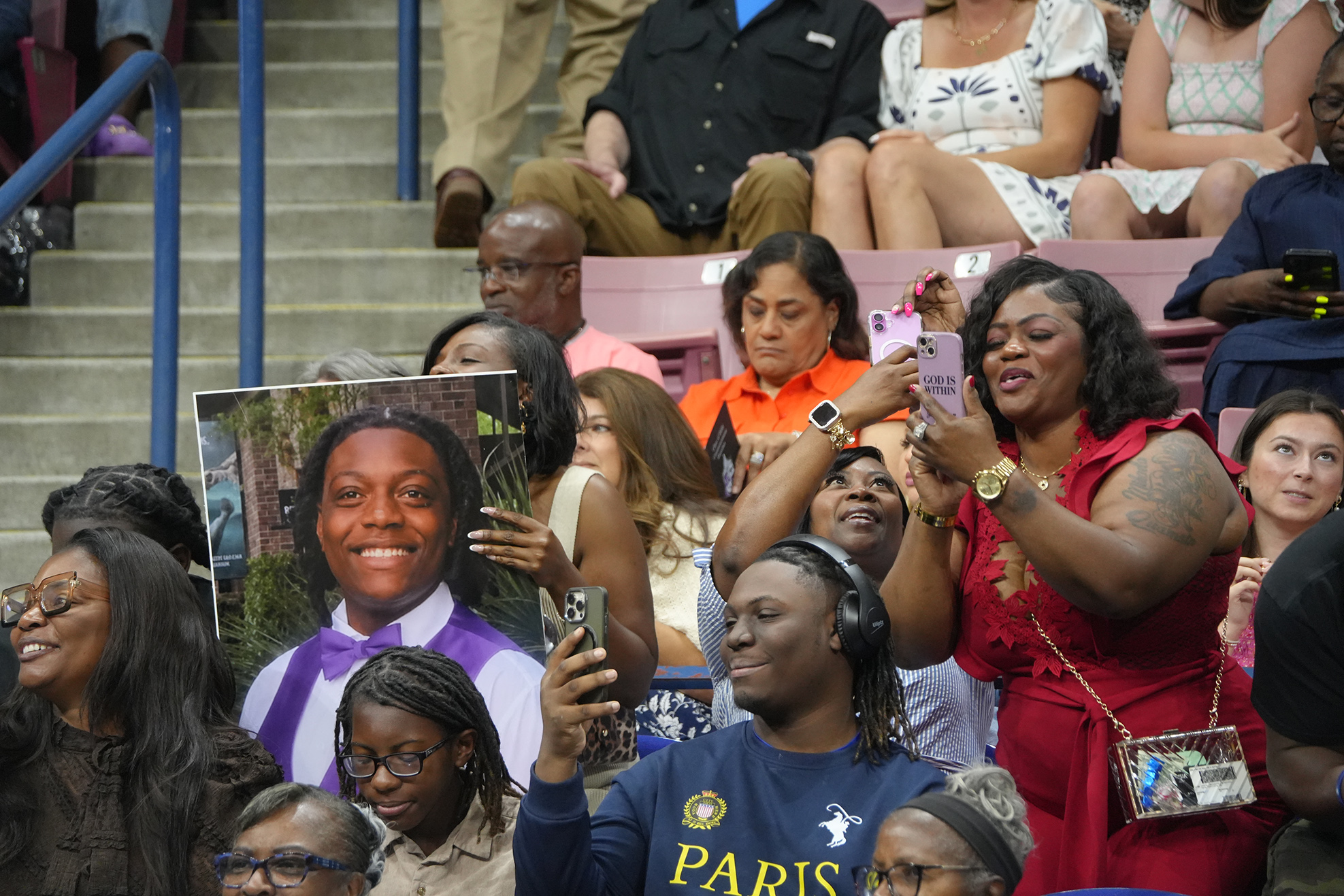 Group of people in a stadium watching an event. Some hold up phones, while others display a poster of a smiling person.