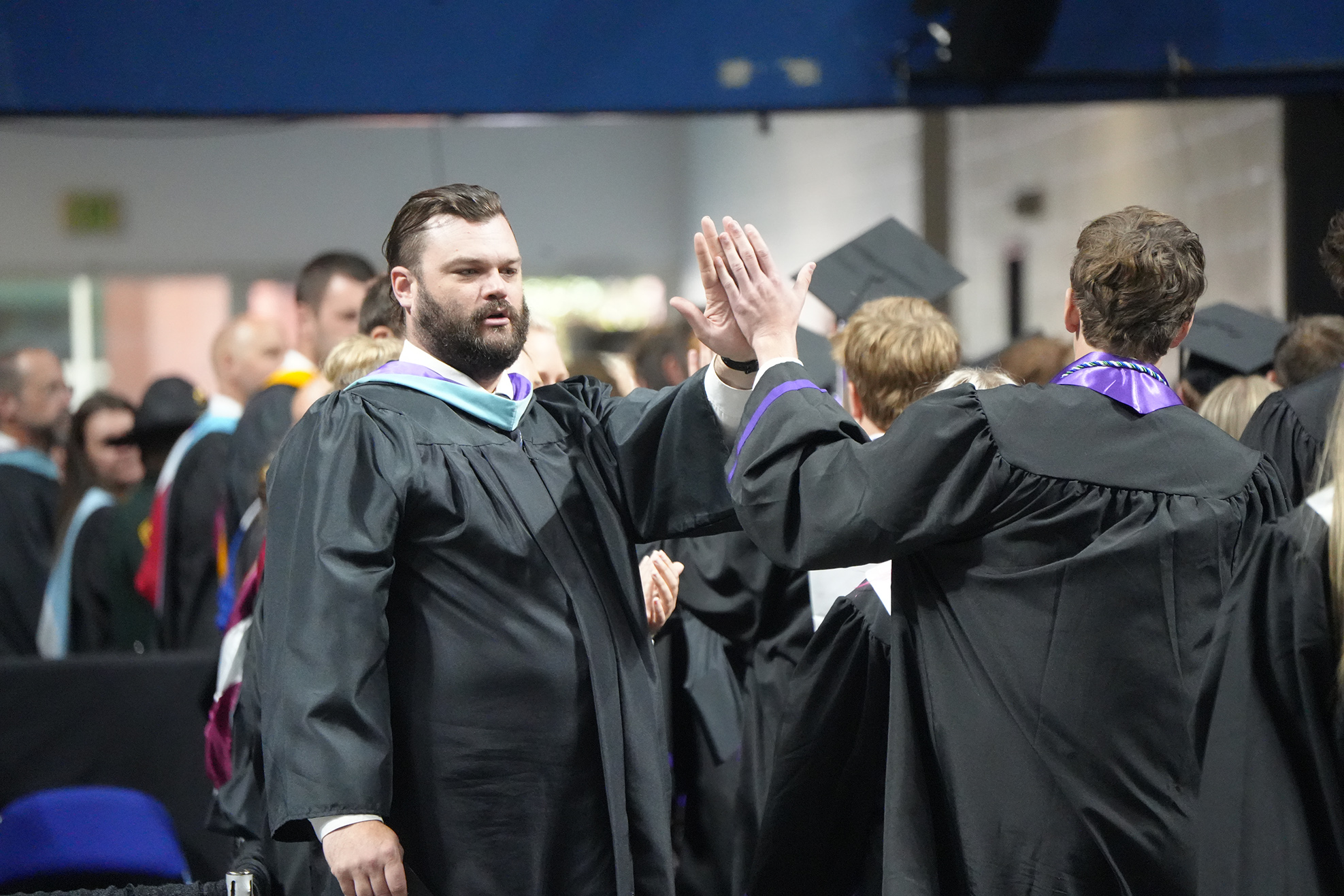 Graduates in black gowns, one giving high five, stand among peers at a ceremony.