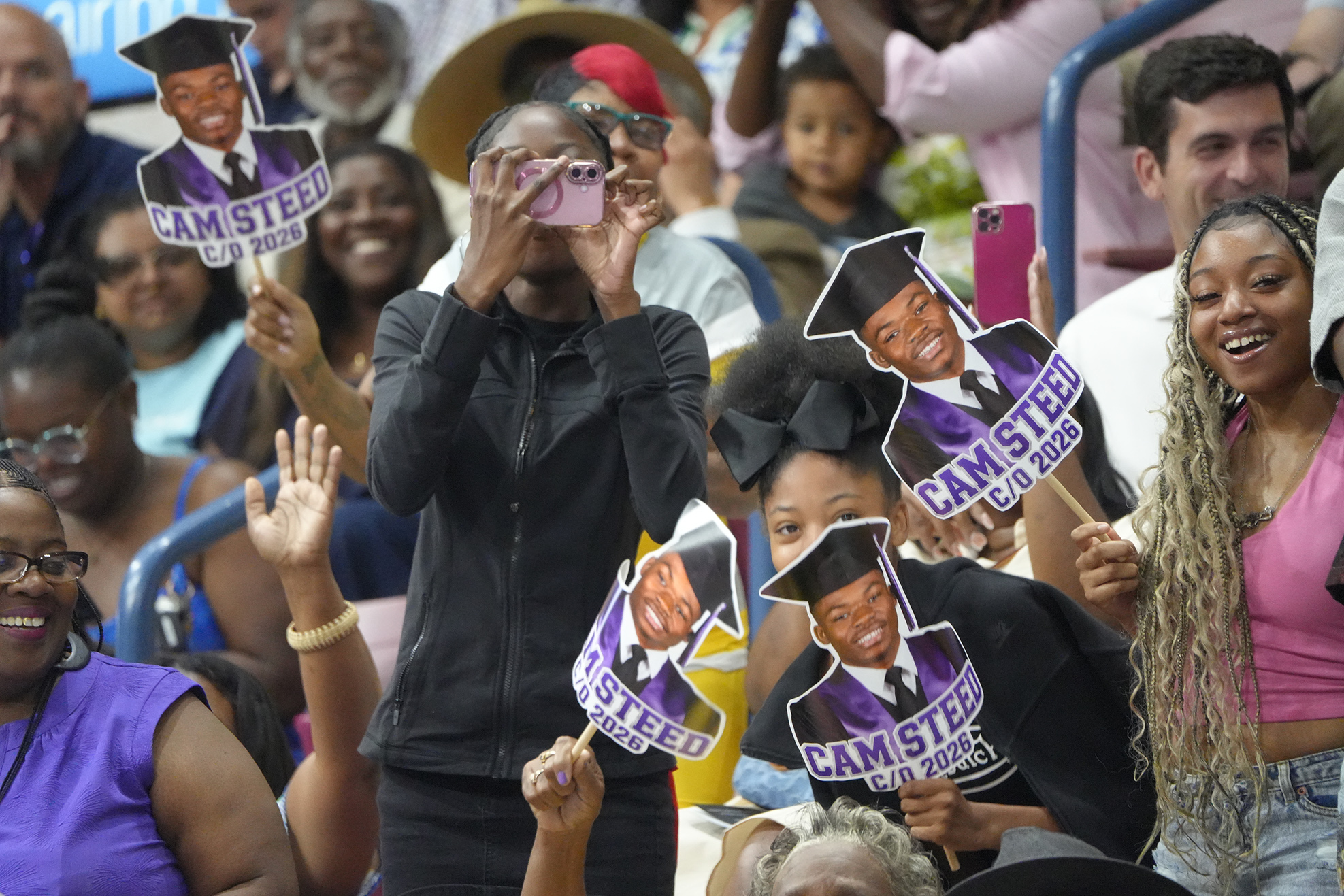 A crowd of people in stands, some wearing graduation caps, hold signs and smile. A person in a black jacket takes a photo.