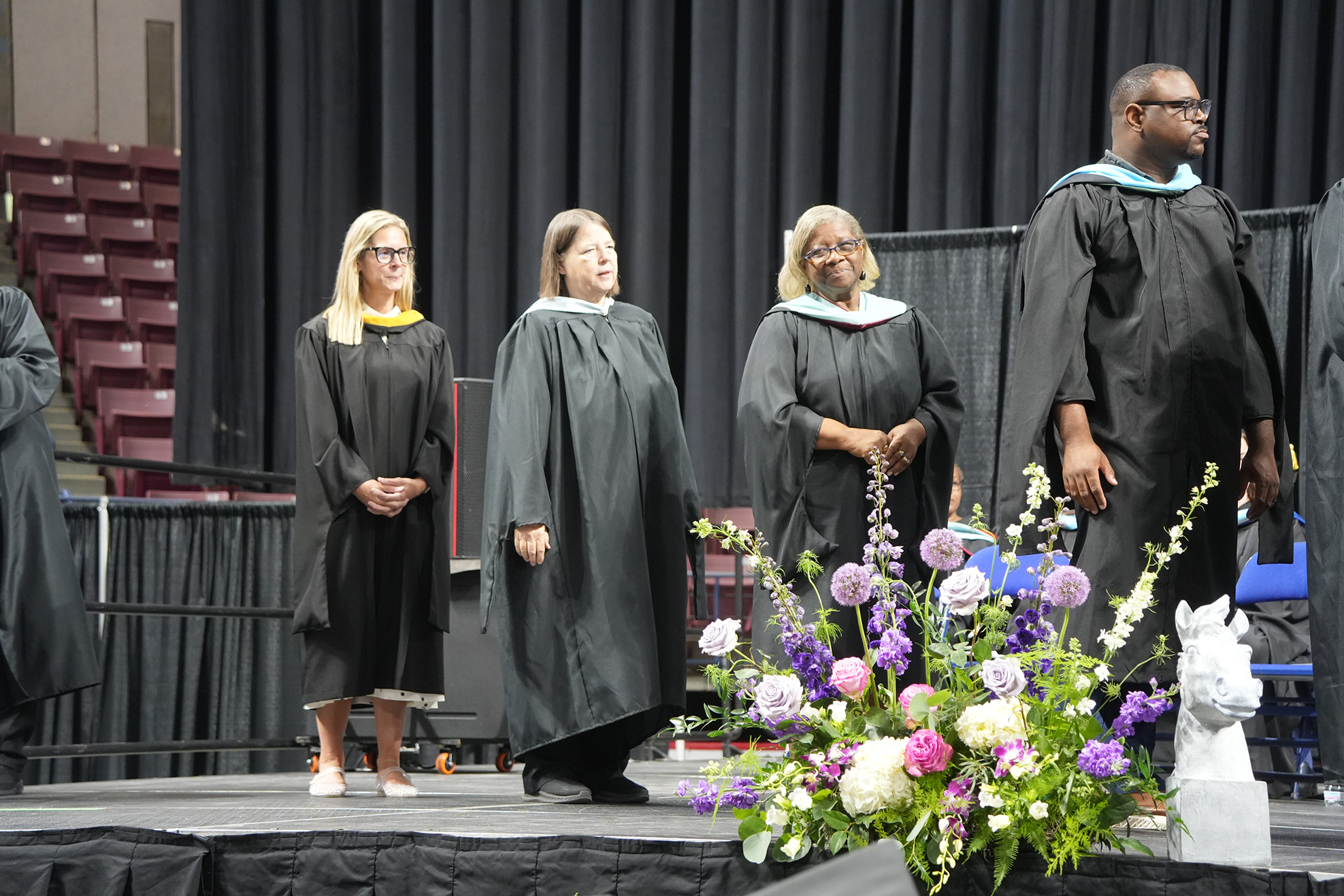 Five individuals dressed in black academic gowns on a stage with flowers in front of them.