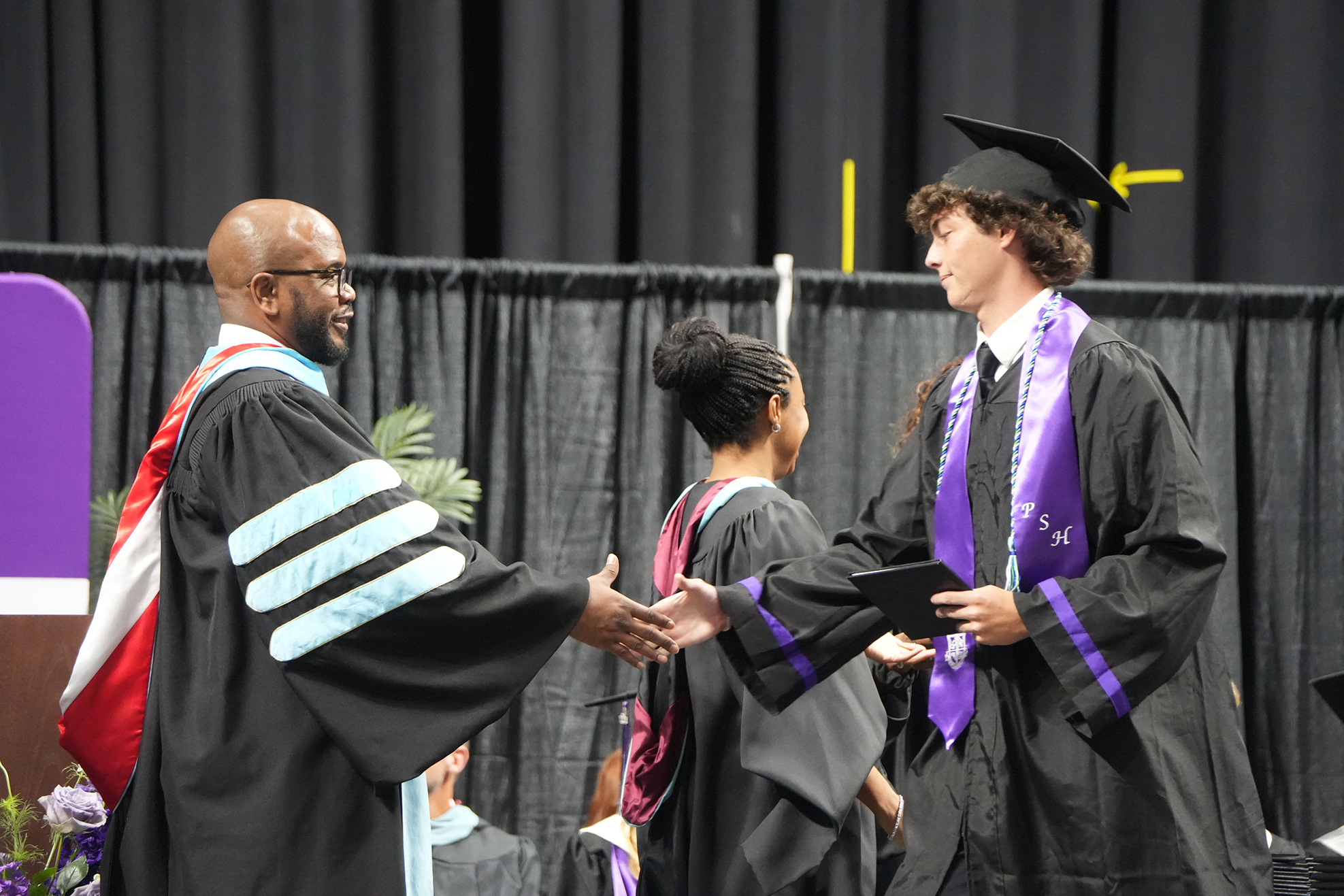Two men in black gowns and caps shake hands. One holds a diploma and wears glasses. Behind them, a woman in a red gown.