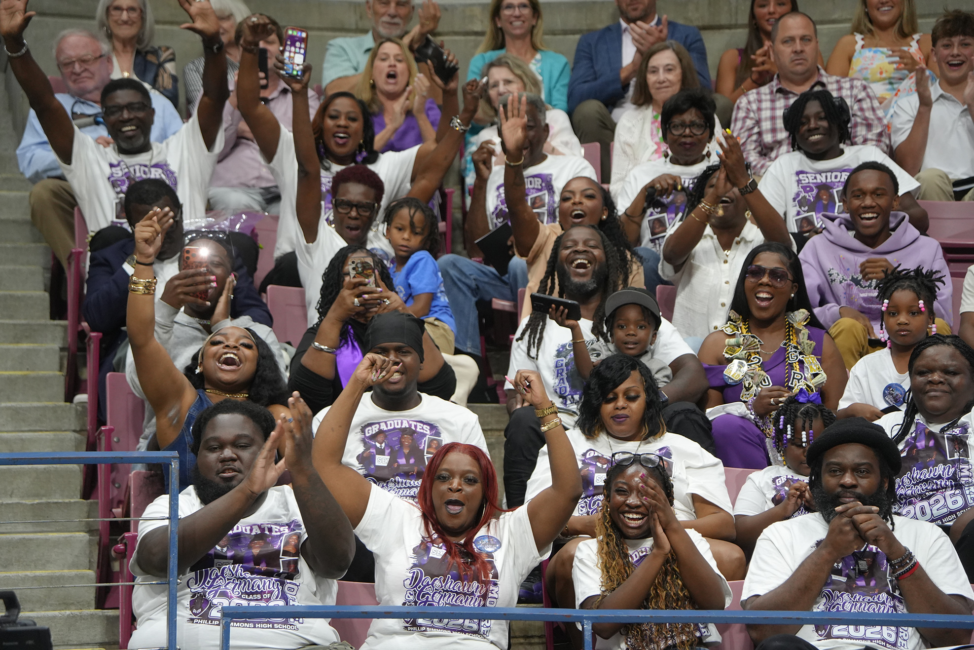 A group of people wearing white t-shirts with purple logos are seated in bleachers, cheering and raising their hands.
