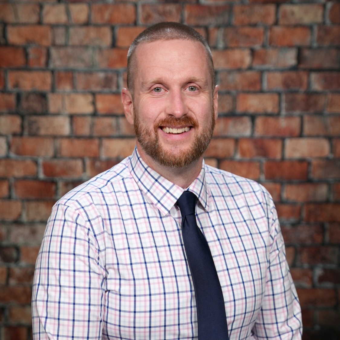 Man in a plaid shirt and navy blue tie standing in front of a brick wall.