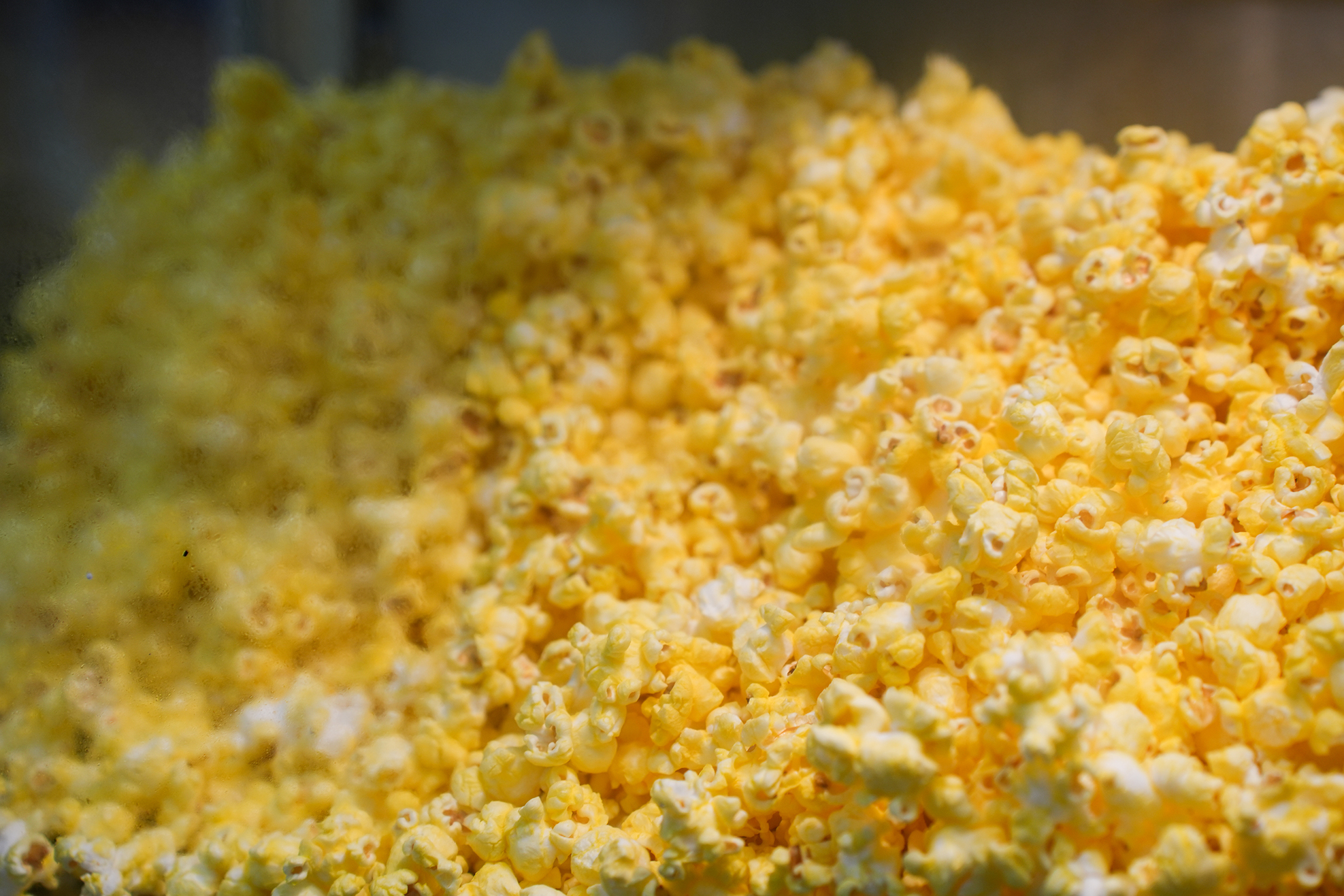 A close-up of yellow popcorn, slightly blurry with a pile in the foreground and a dark background.