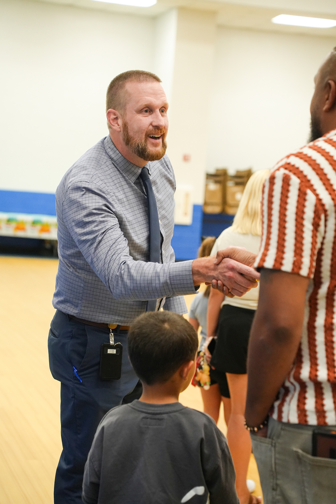 A man in a light blue shirt and tie shakes hands with another man in a striped shirt. Behind them, a child and a woman in a black skirt are present.