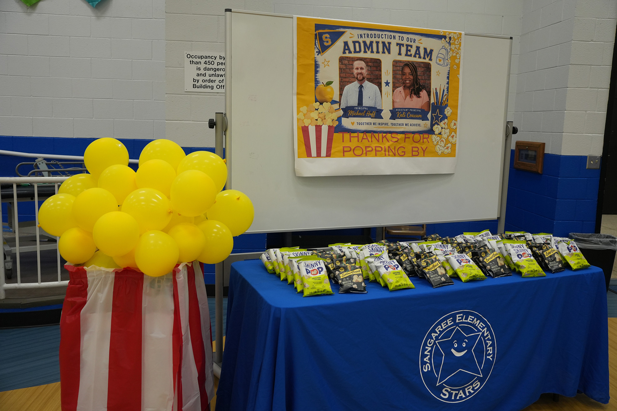 Yellow balloons and popcorn bags on a table next to a banner. A whiteboard and sign behind it.