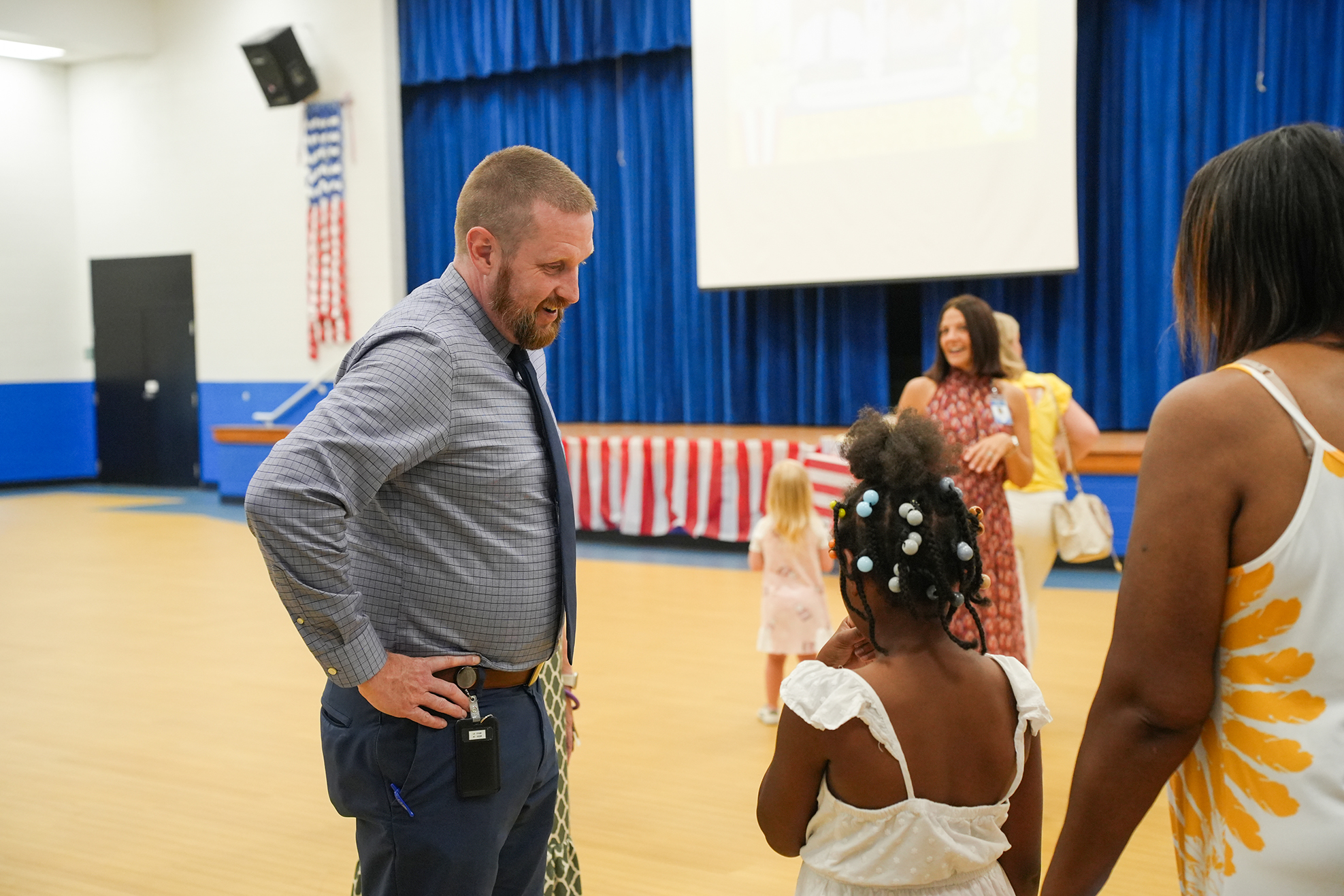 A man and two women, one in a tie, stand next to a young girl in a white dress. Behind them, a woman walks with a small child.