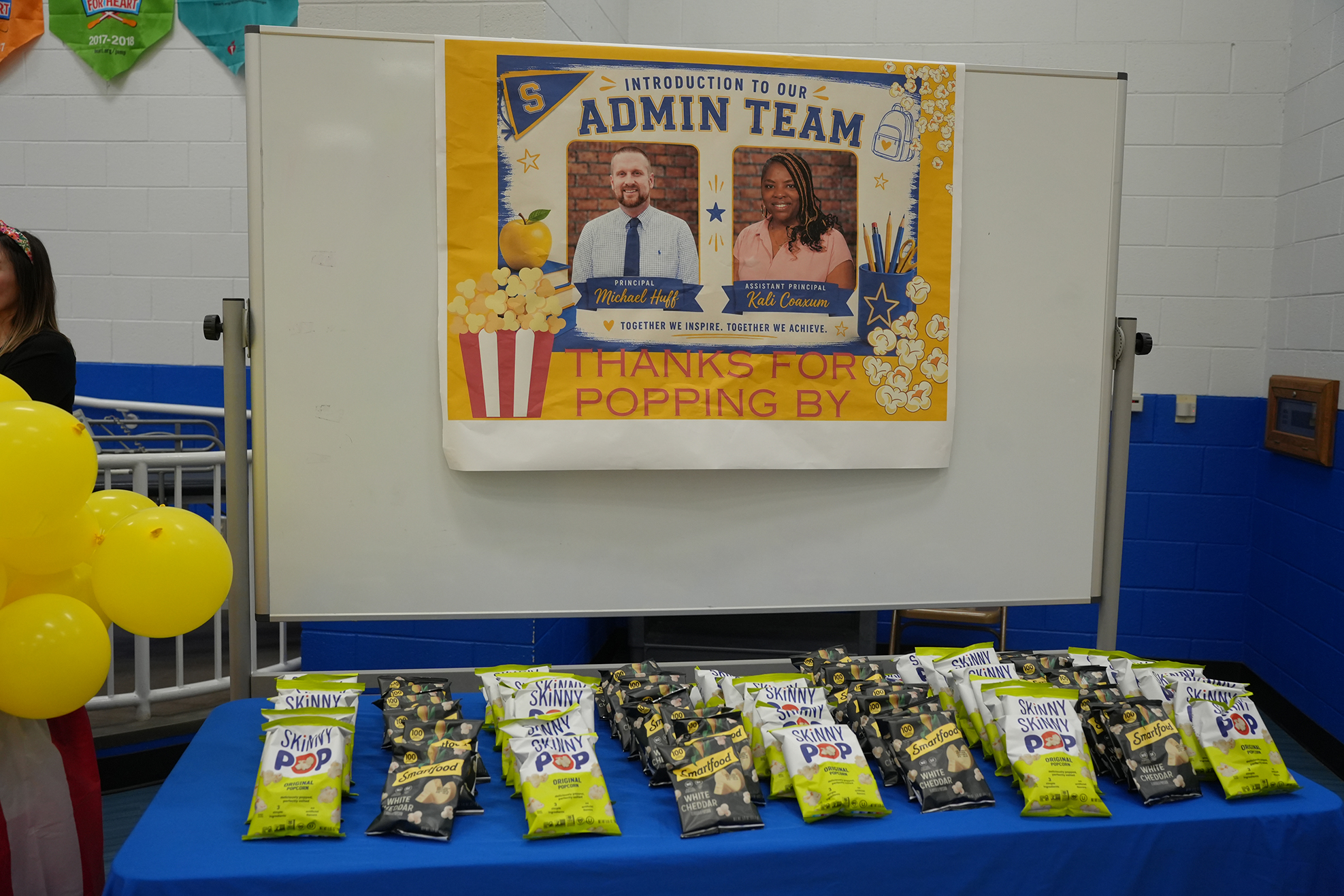 A white board with a banner featuring two people and a message. Below it, a table displays multiple bags of popcorn.