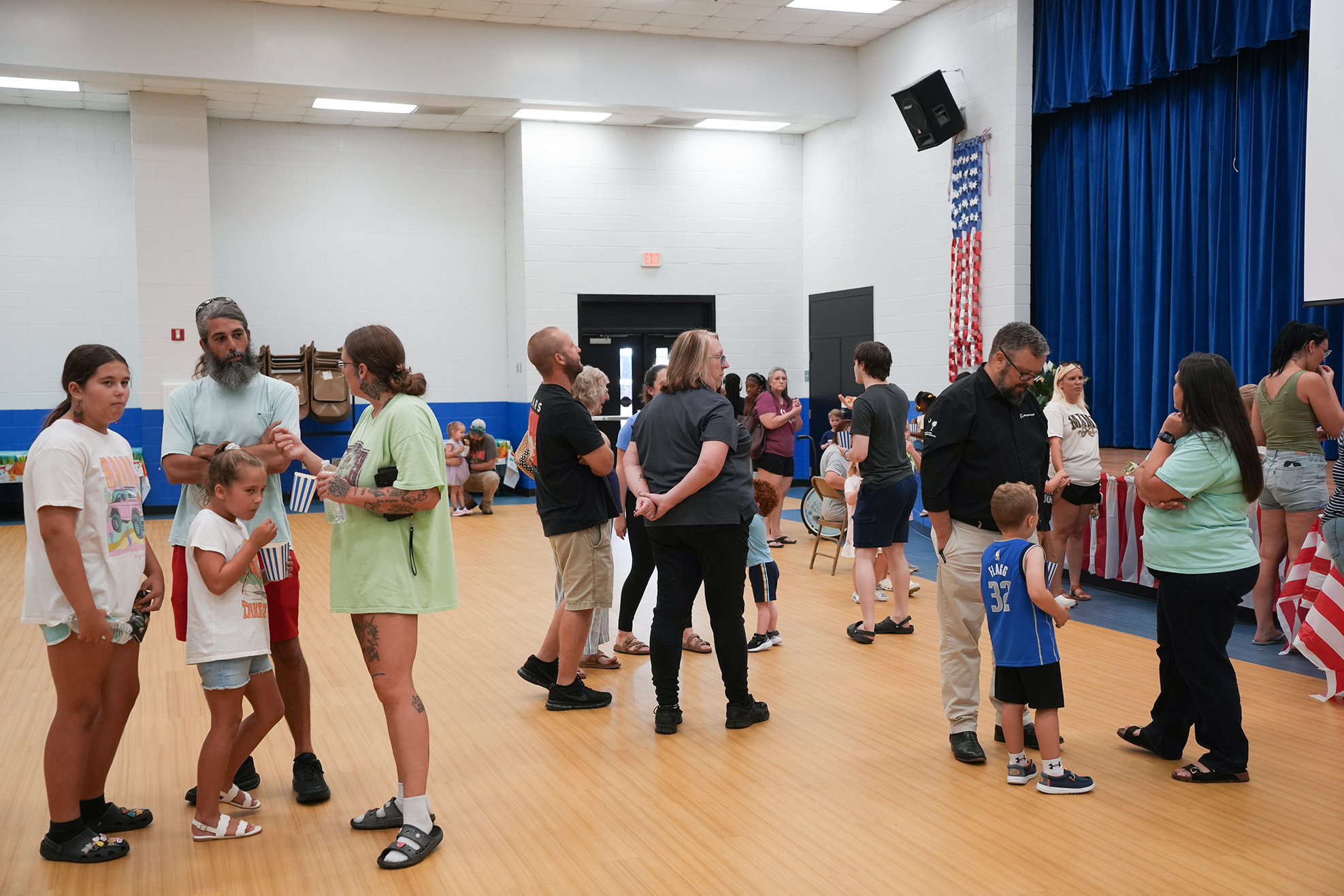 Several people, including adults and children, are in a room with wooden flooring and blue curtains.