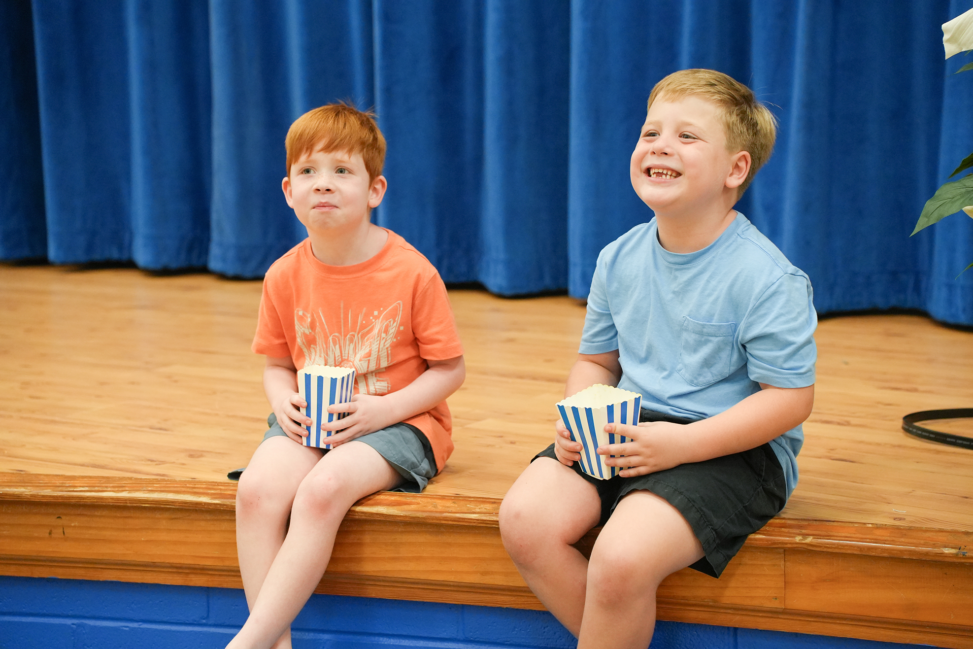 Two children sit on a raised platform, each holding a cup, smiling at the camera. A blue curtain and a blue floor are in the background.