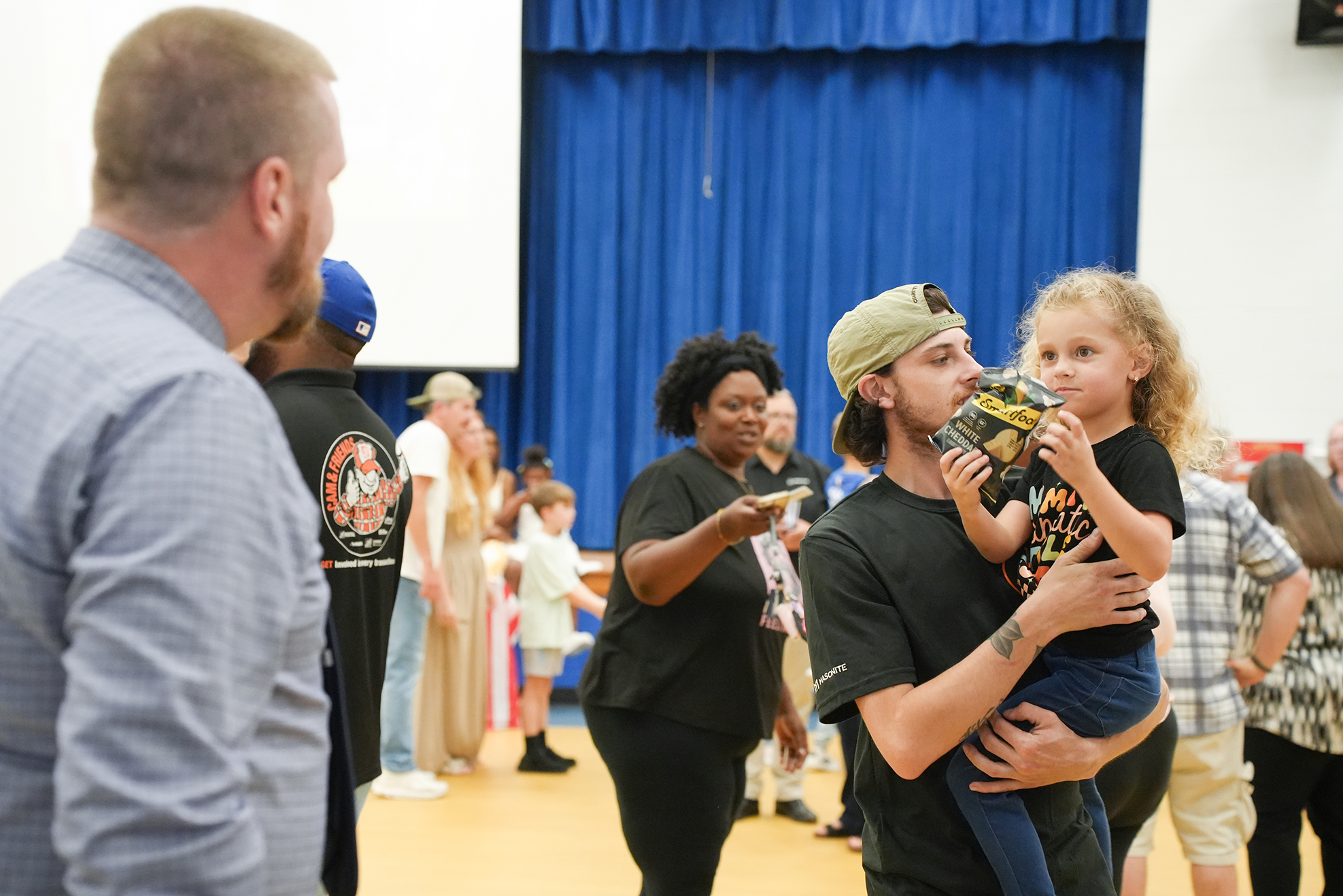 A group of adults and children in a room with blue curtains. One man holds a child while another adult faces him.